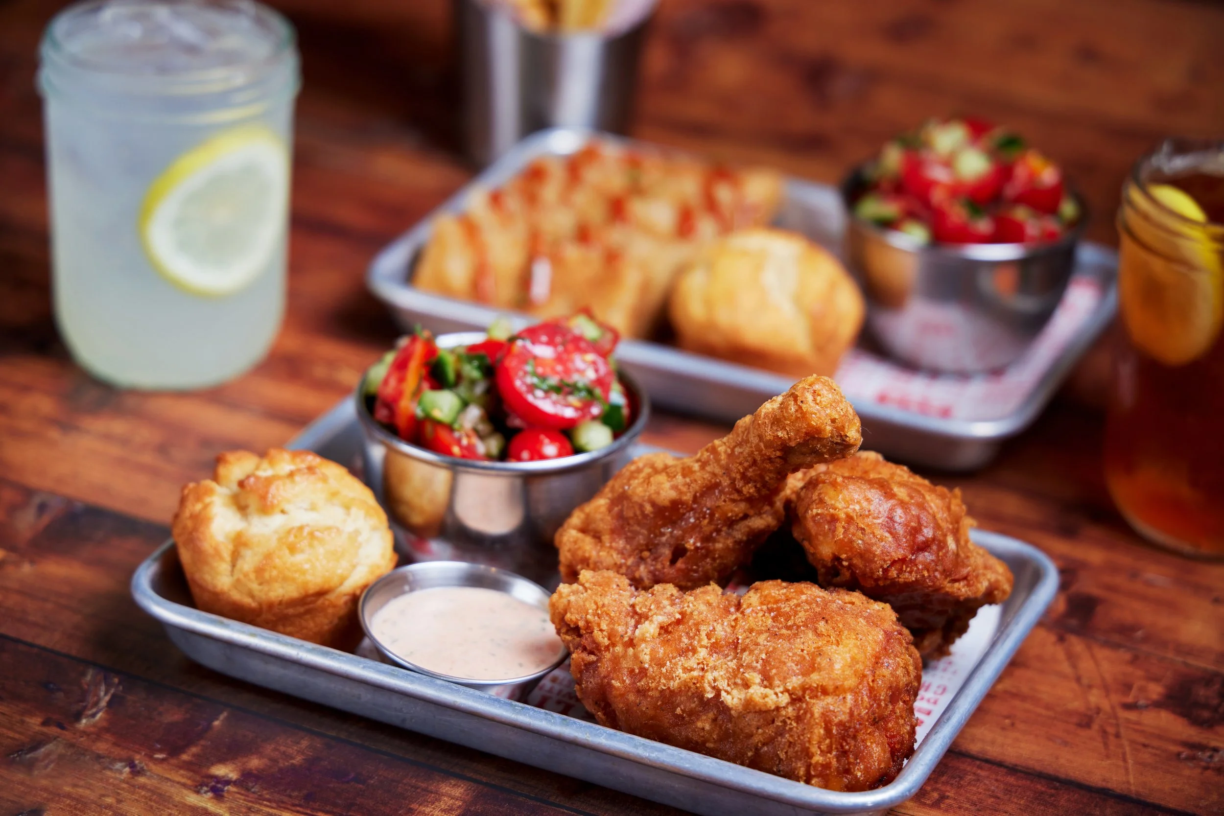 A tray of fried chicken, small bowls of salad and dipping sauces, and a drink with lemon slices on a wooden table.