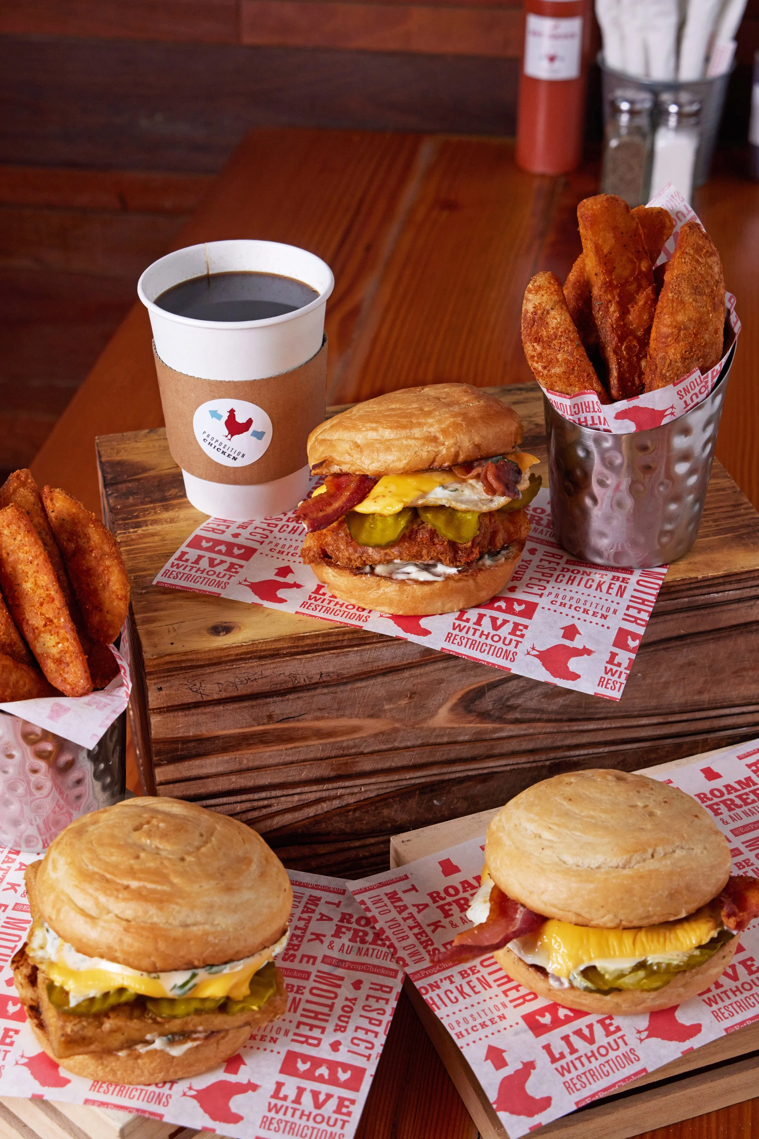 A wooden tray holding a sandwich with fried chicken, egg, and cheddar cheese, a cup of black coffee in a paper cup, and a cup of seasoned french fries in a metal container. Other similar sandwiches and buns are visible on a wooden table.