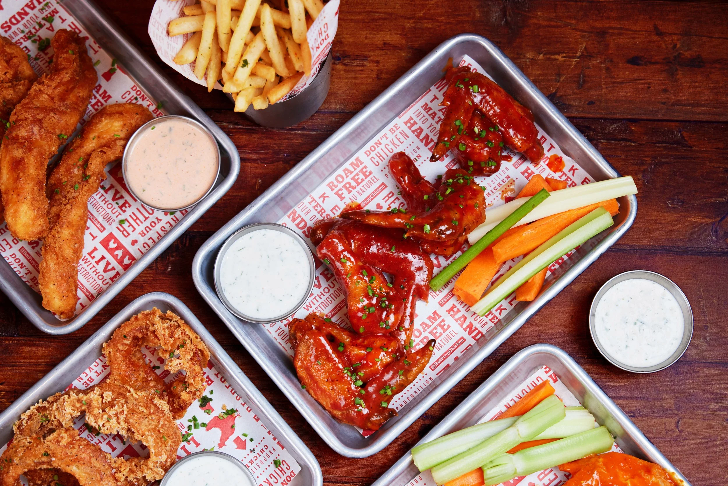A variety of fried chicken strips, French fries, buffalo chicken wings, and fresh vegetable sticks served with multiple dipping sauces on metal trays lined with branded paper.