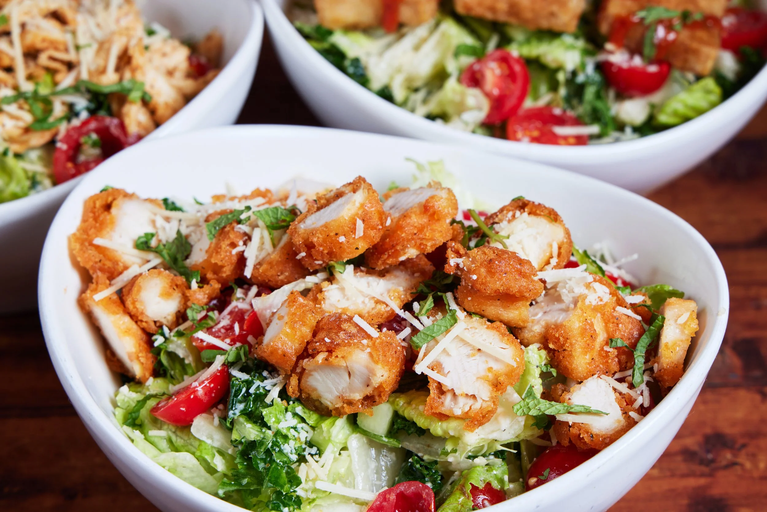 Bowl of chopped fried chicken on a bed of lettuce, shredded cheese, and tomatoes, with other bowls of salad nearby.