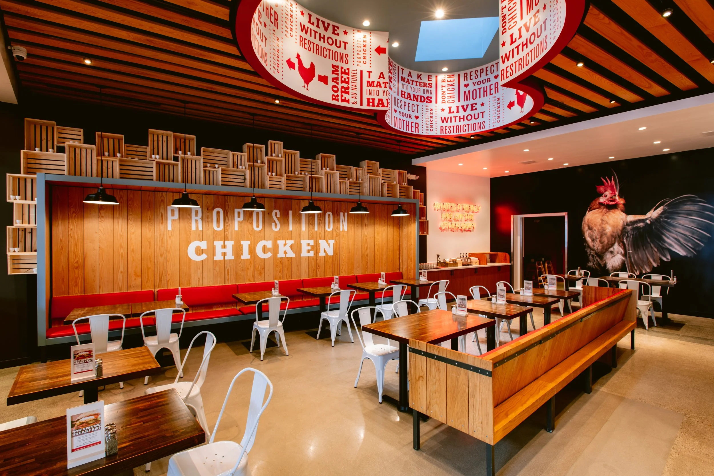 Interior of Propagation Chicken, featuring wooden tables and chairs, a wooden wall with a white sign, and a decorative rooster mural on a black wall. The ceiling has wooden slats with hanging industrial lights.