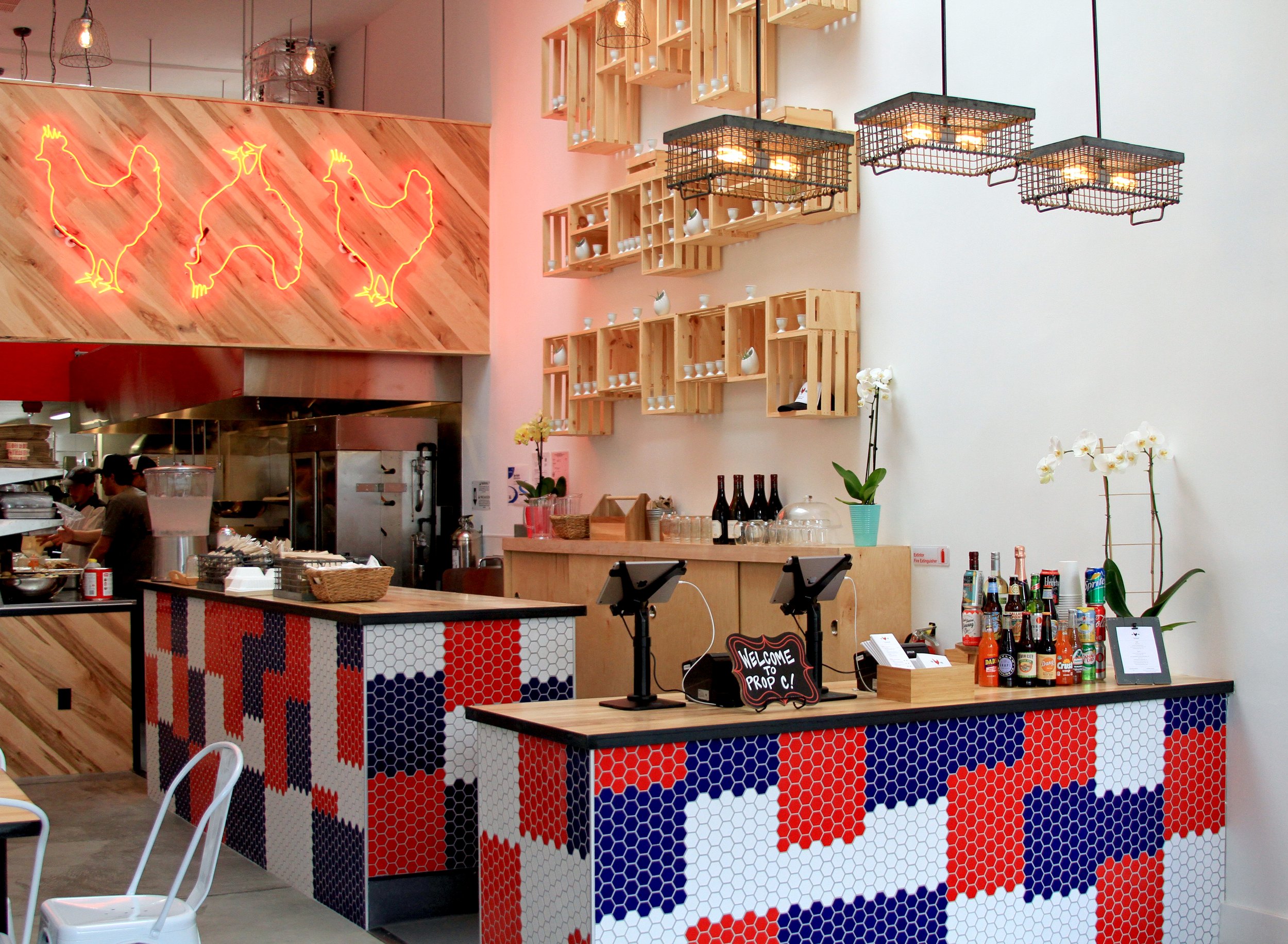 Interior of Proposition Chicken in Oakland with wooden and hexagon tile decor, hanging grid light fixtures, a colorful patterned bar front, black chairs, tables with bottles and condiments, and a wall with wooden shelving and a neon rooster sign.