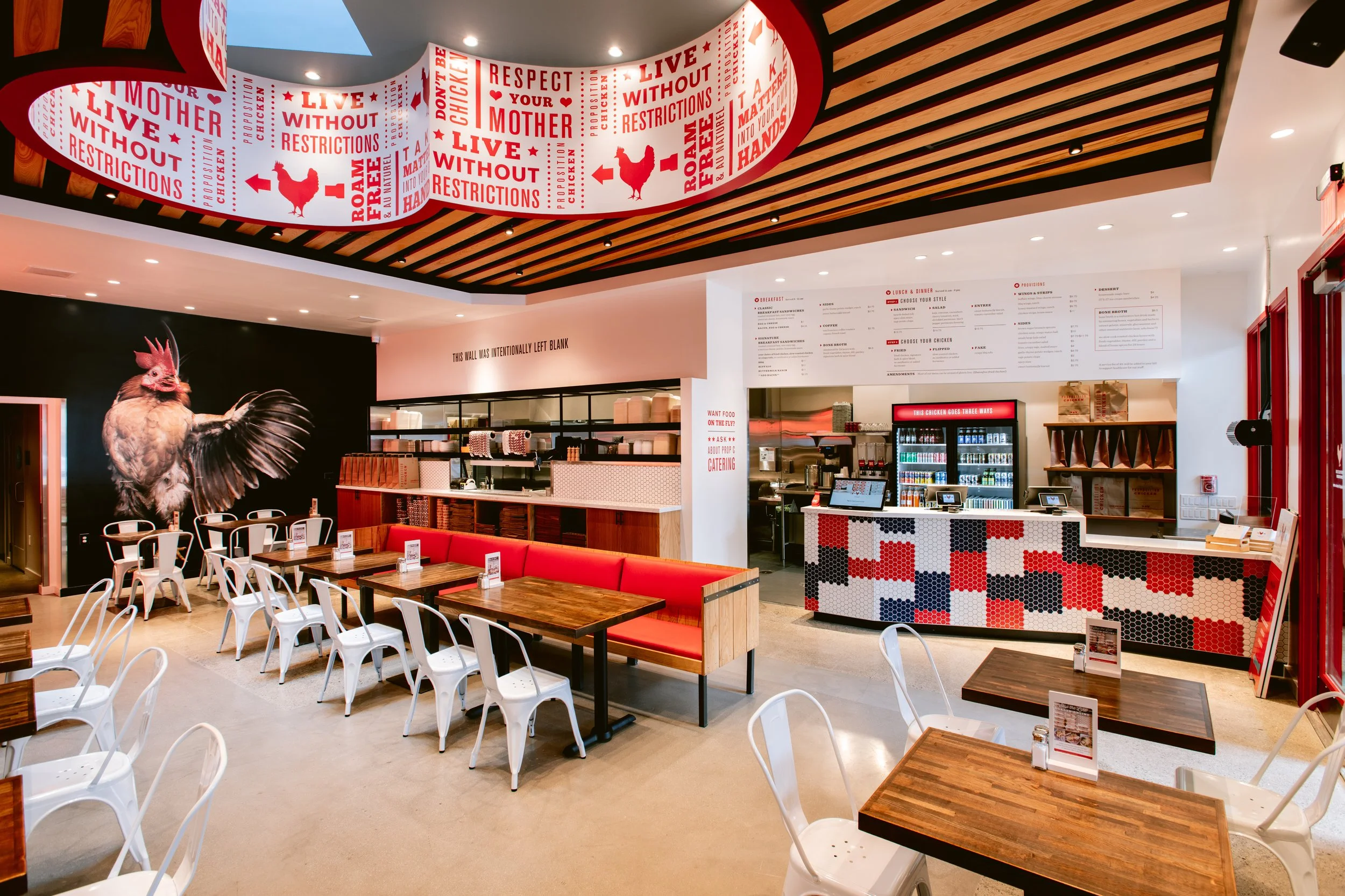 Interior of Proposition Chicken San Francisco with white chairs, wooden tables, red cushioned benches, a mural of a rooster on a black wall, and a ceiling with wooden slats.