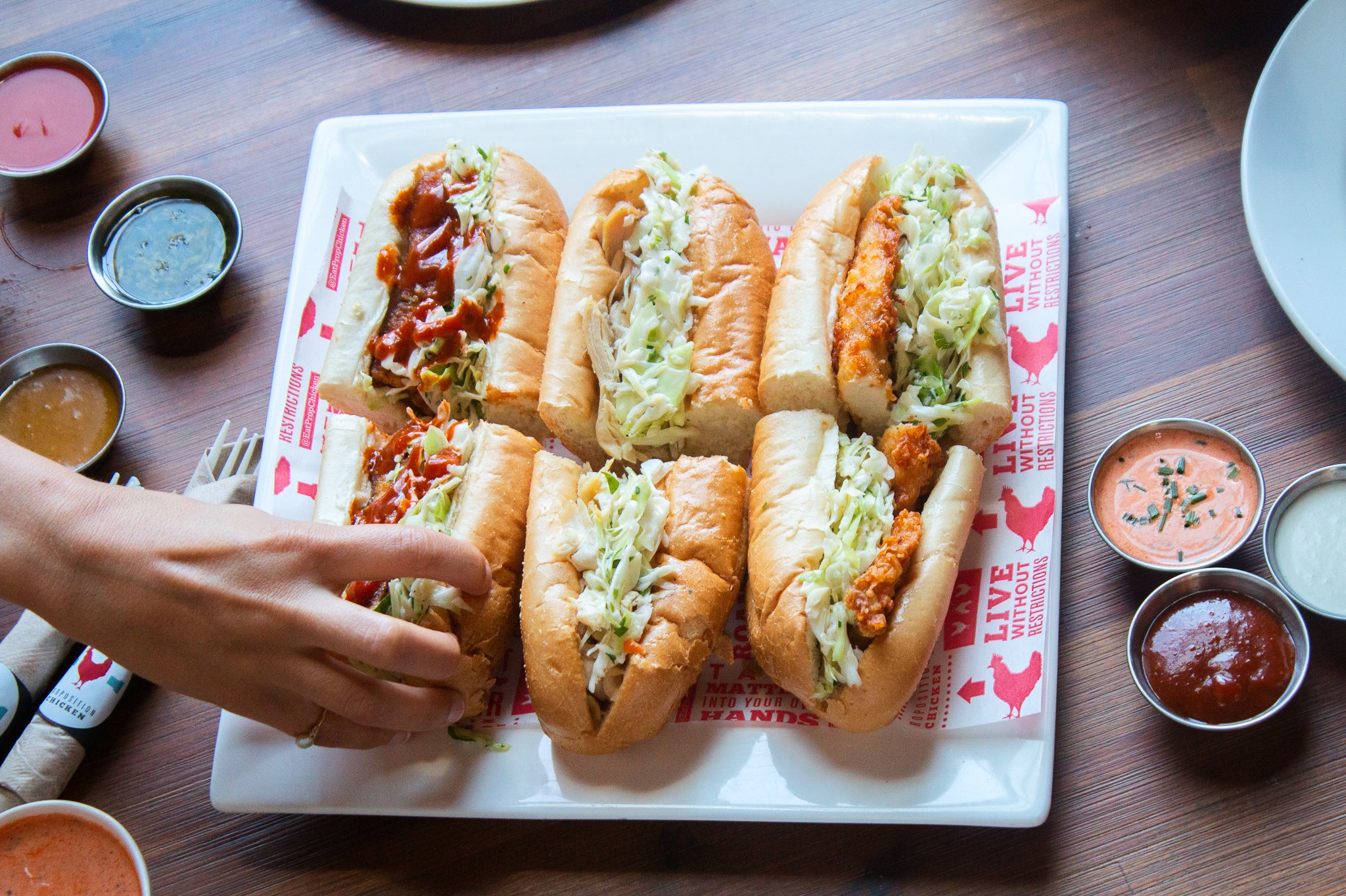 A tray of five sandwiches with shredded lettuce and various fillings, surrounded by small bowls of different sauces on a wooden table.