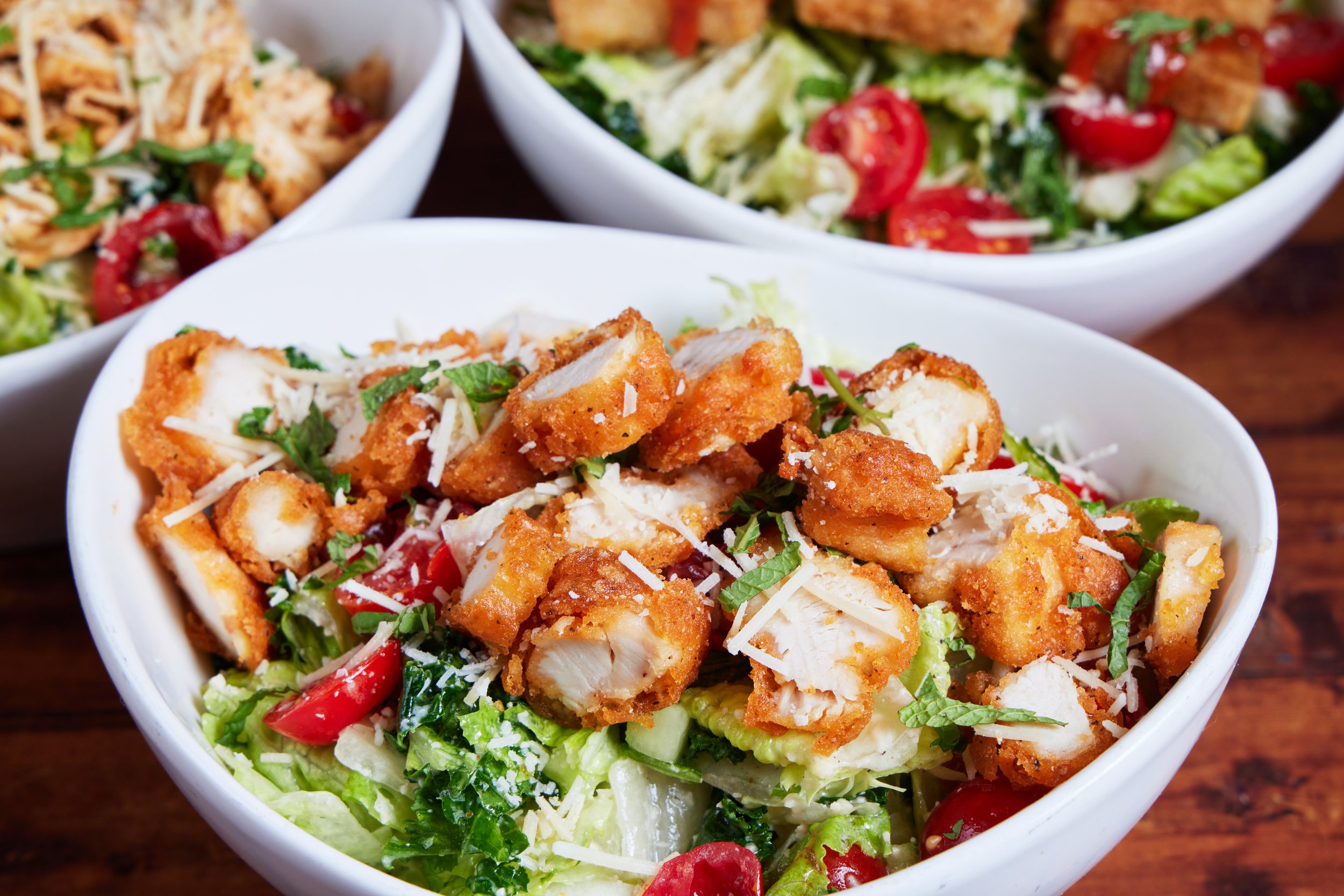 Bowl of chopped fried chicken on a bed of lettuce, cherry tomatoes, shredded cheese, and herbs, with two more bowls of salad in the background.