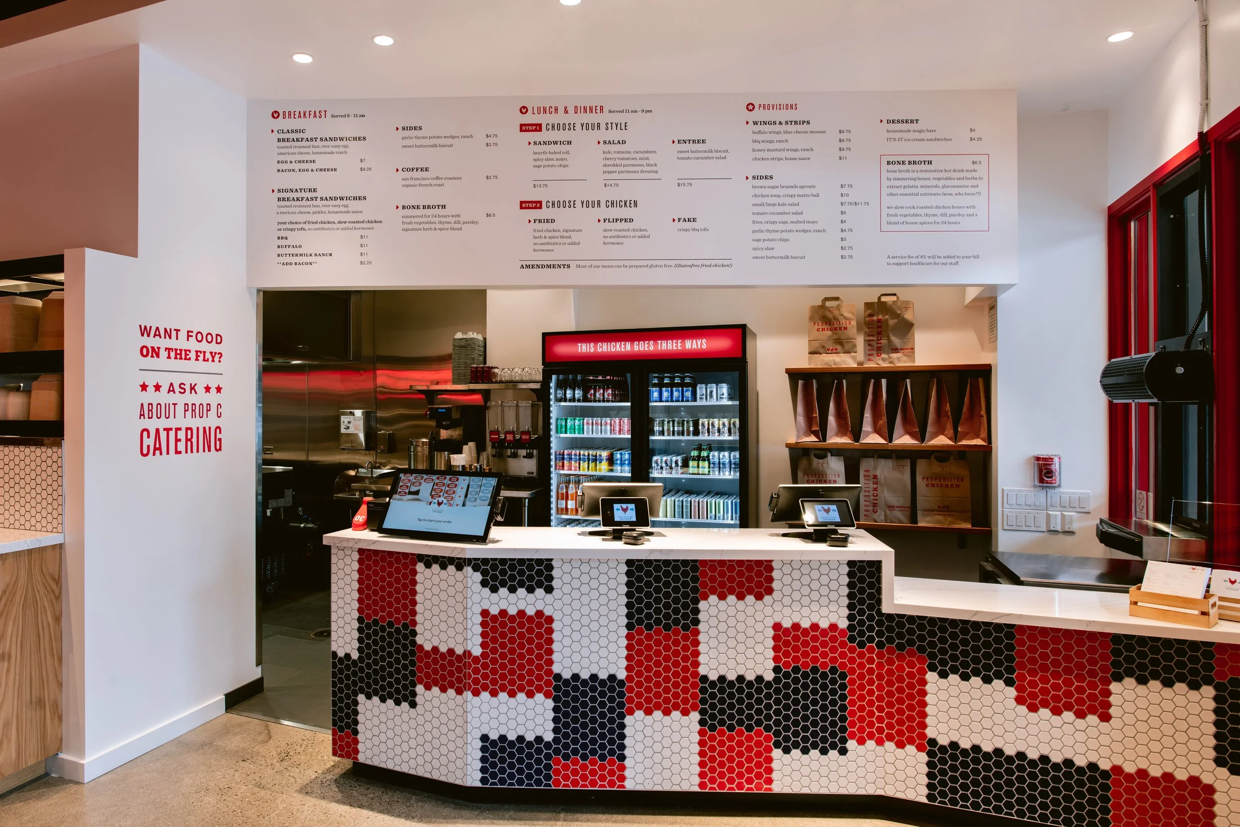 Ordering counter inside Proposition Chicken San Francisco with a red, black, white, and gray hexagonal pattern, digital order screens, refrigerator with drinks, and a menu on the wall behind. 