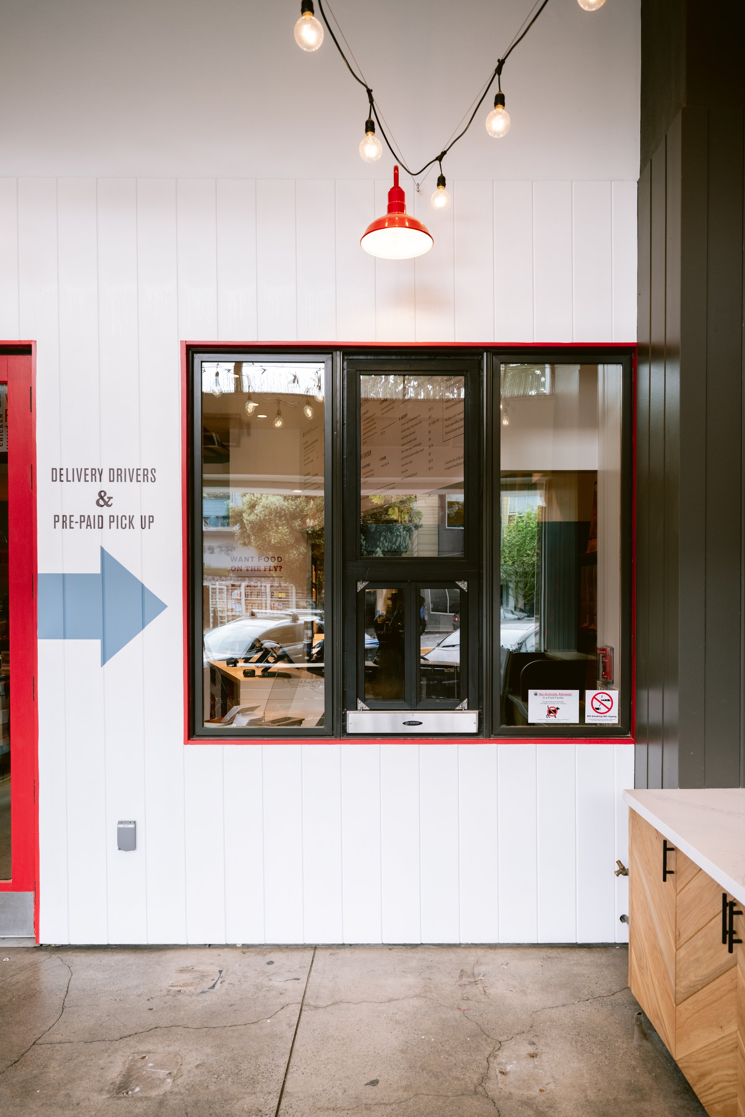 Inside view of food pickup window at Proposition Chicken. Signs indicating delivery and pickup options. Part of a wooden counter is seen in the foreground.
