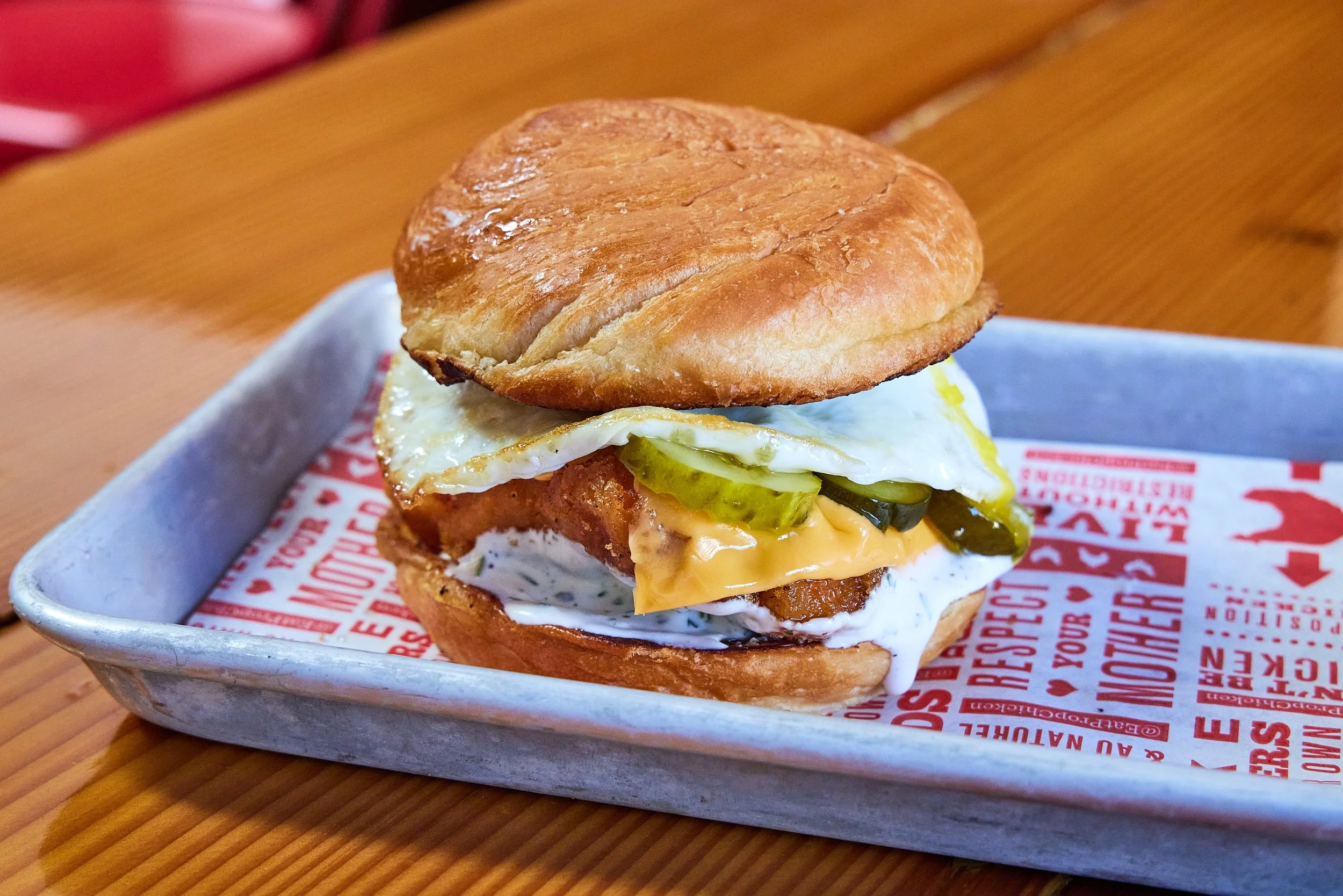 A breakfast sandwich on a toasted biscuit, with fried egg, pickles, melted cheese, and chicken, served on a paper-lined tray.