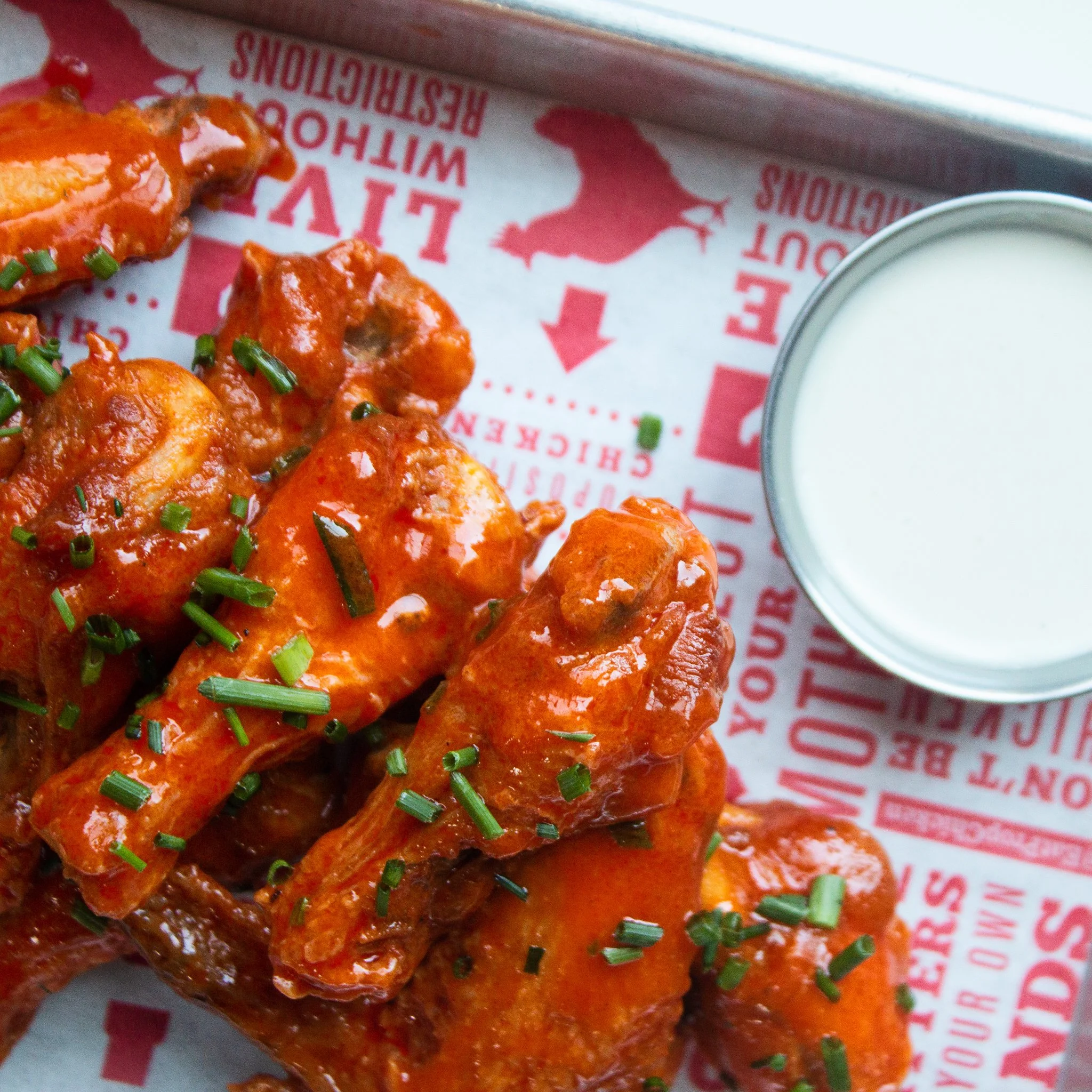 Close-up of chicken wings topped with green chopped chives on red and white paper branding, next to a small container of white dipping sauce.