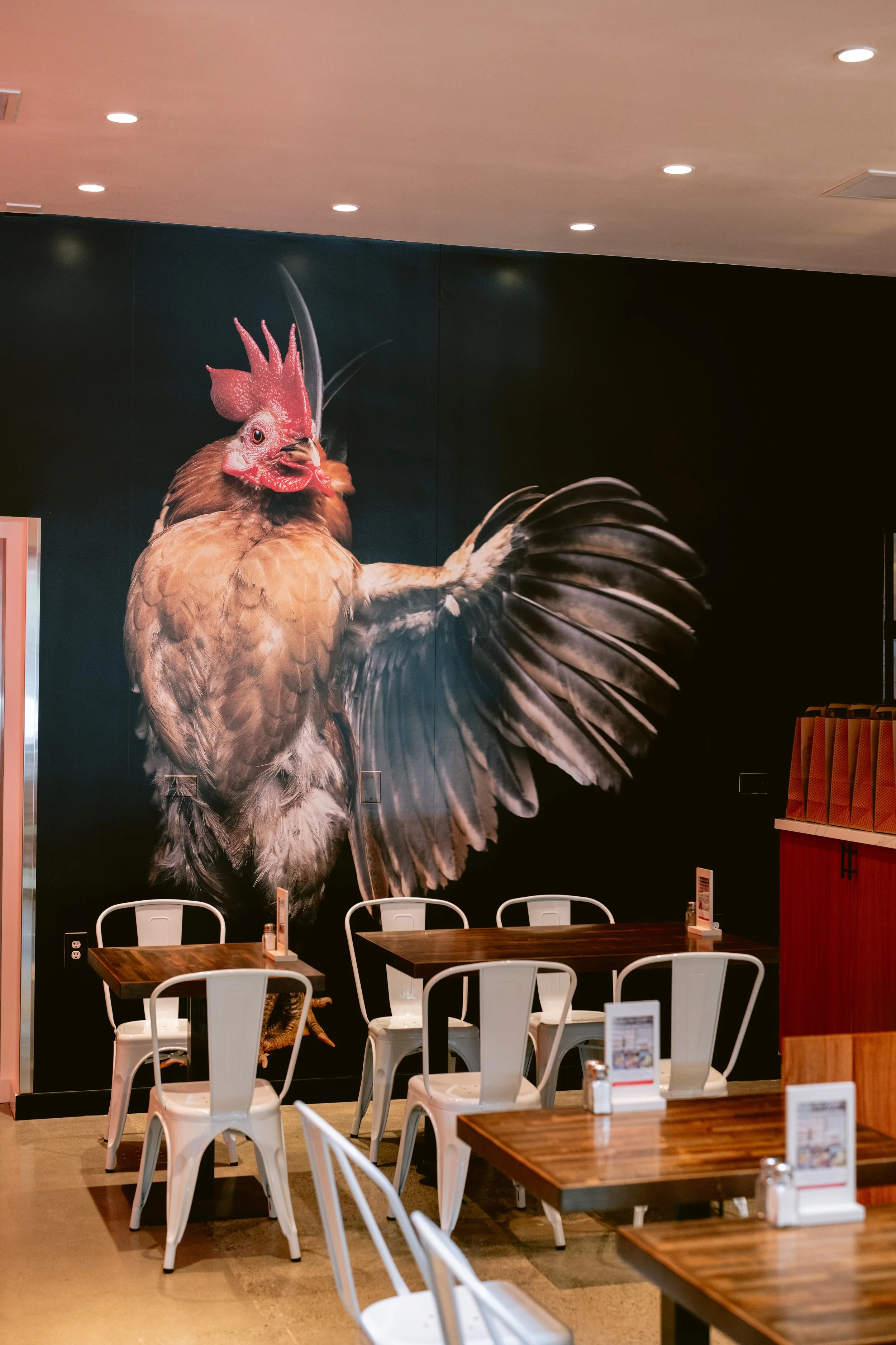 Interior of Proposition Chicken San Francisco, with a large wall mural of a rooster, sitting at tables with white chairs, menu stands on tables.