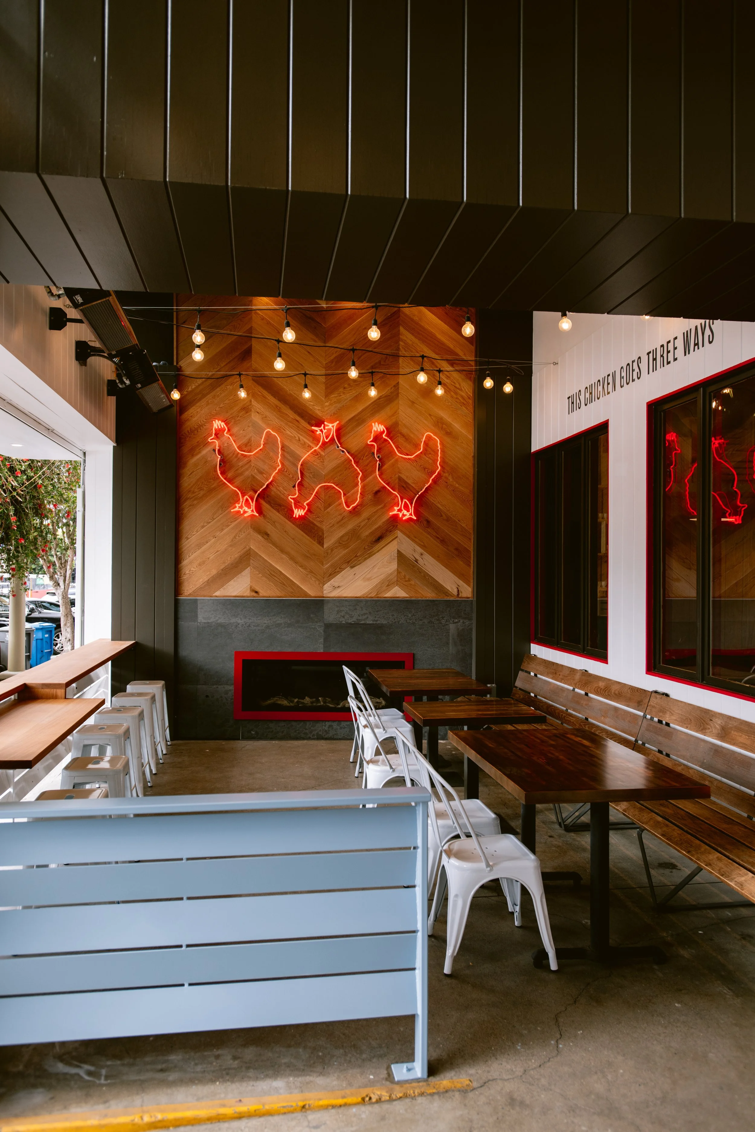 Patio seating area at Proposition Chicken San Francisco with wooden tables, white and metal chairs, decorative neon chicken sign, and string lights on a wooden wall.