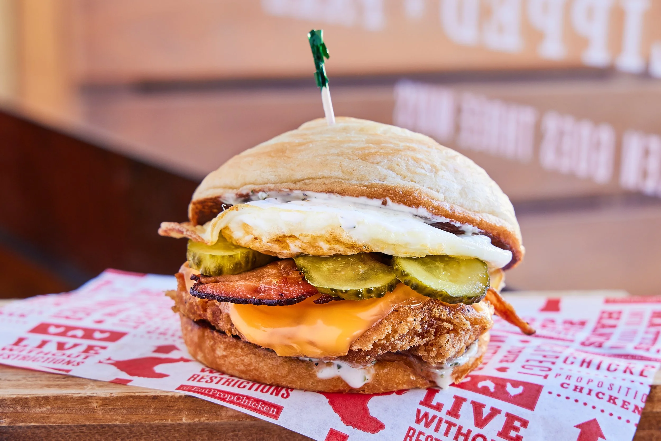 A close-up of a layered breakfast sandwich with pickles, bacon, cheese, a fried chicken patty, a sunny side-up egg, and creamy sauce, topped with a toasted bun.