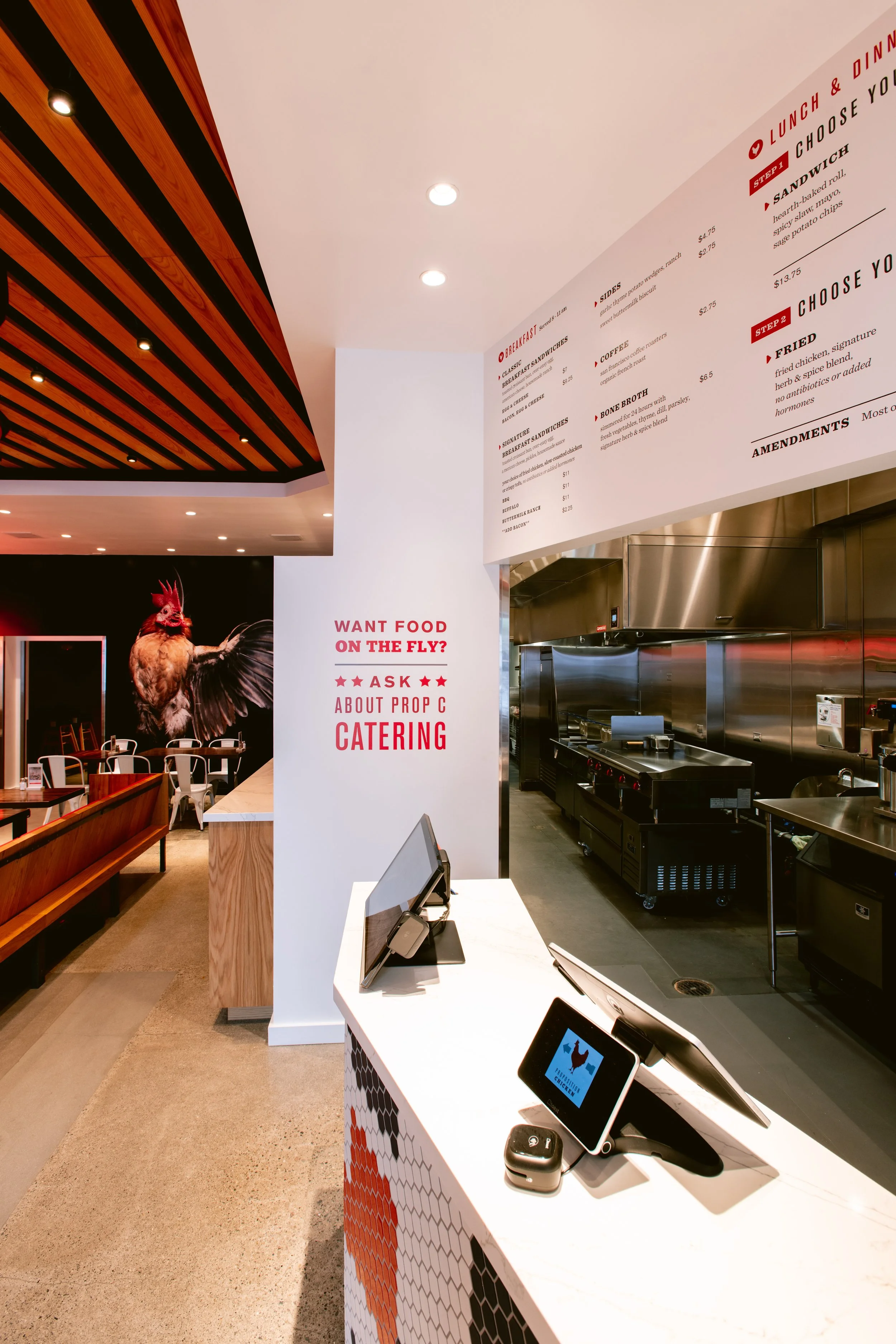 View of Proposition Chicken ordering counter with ordering tablets, a stainless steel kitchen area, and a decorative wall featuring a rooster mural. The interior has a modern design with wood paneling on the ceiling.