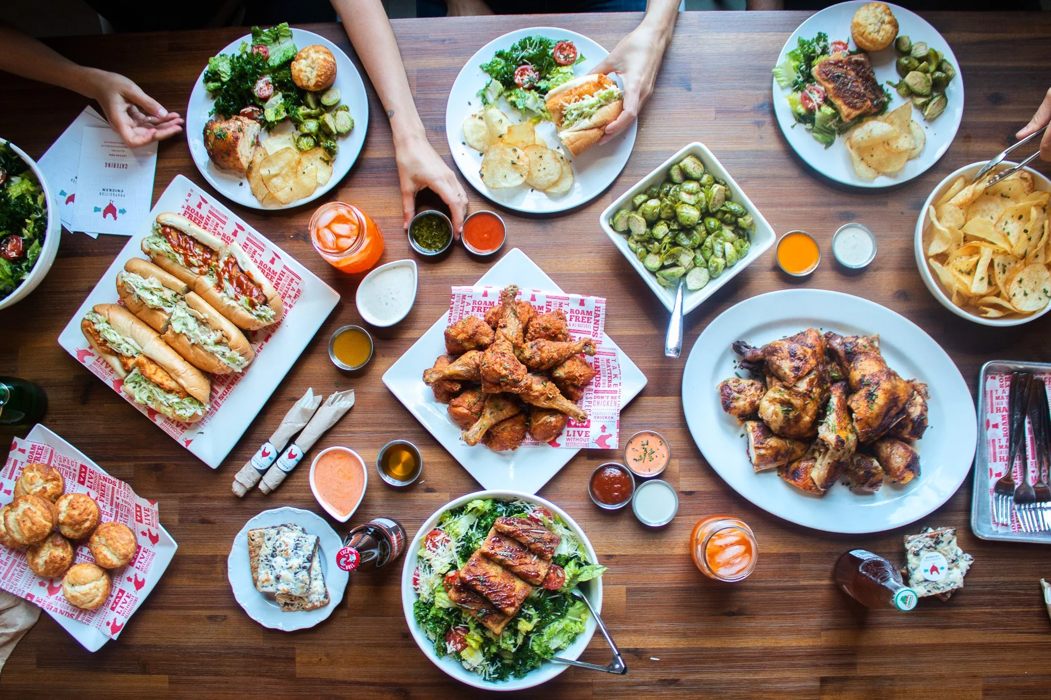 A top-down view of a large brown wooden table filled with plates of food. There are various salads, fried chicken, sandwiches, potato chips, biscuits, and multiple dipping sauces and drinks.