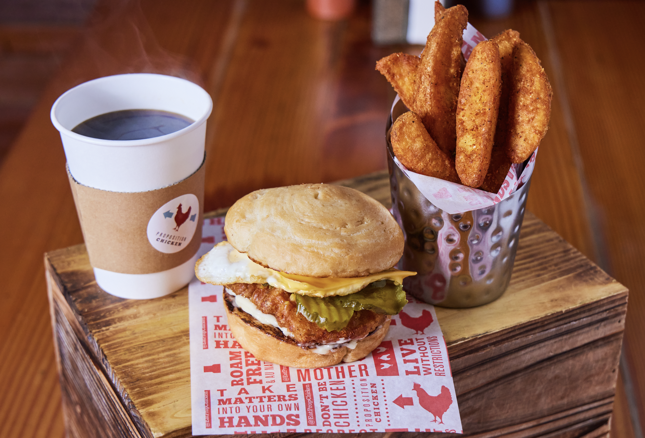A wooden tray holds a paper cup of black coffee, a breakfast sandwich, and a metal cup filled with seasoned potato wedges.
