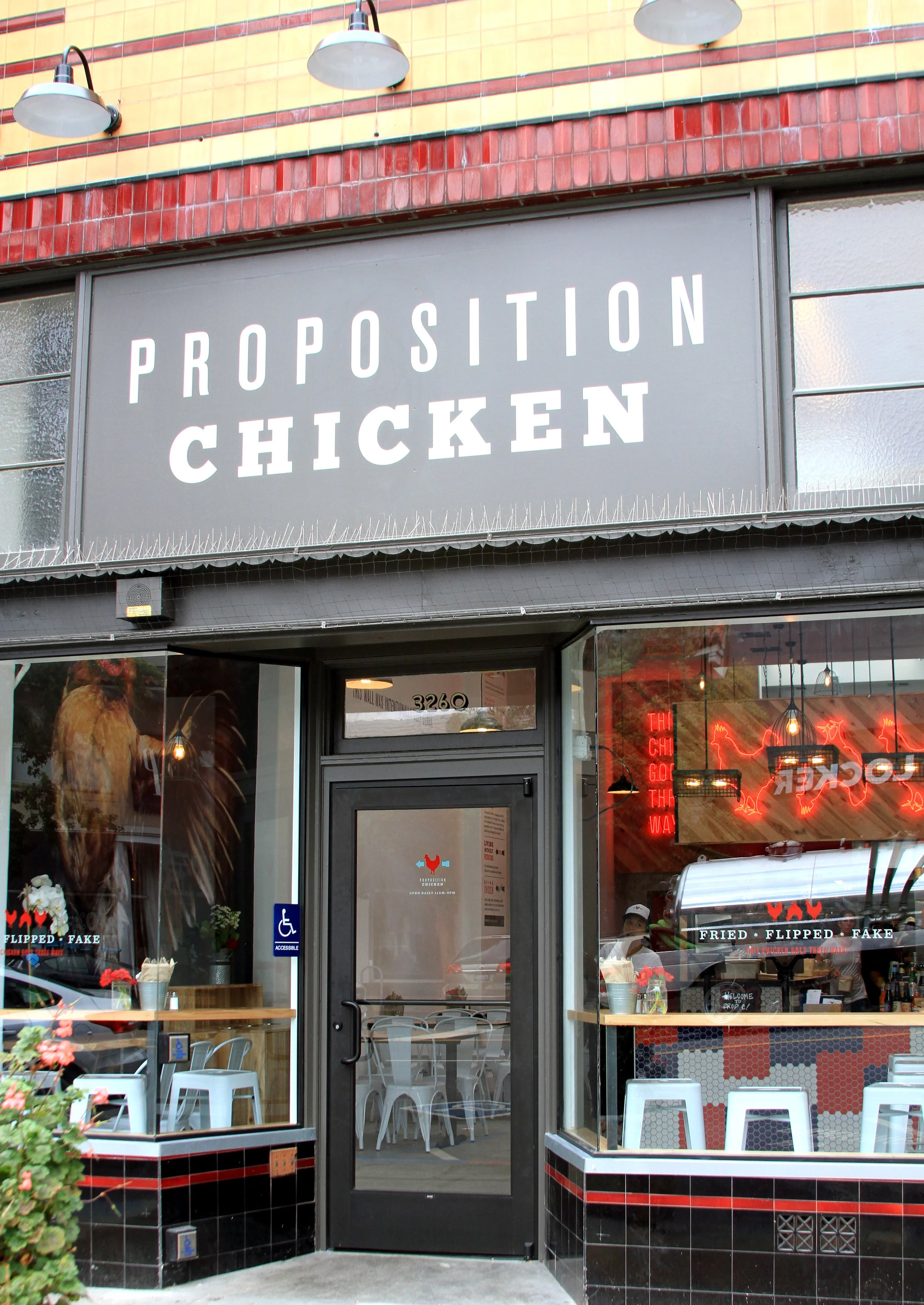 Exterior of Proposition Chicken in Oakland with a large gray sign, glass windows, and a door with the restaurant's logo. Inside, visible seating and neon signs can be seen through the windows.