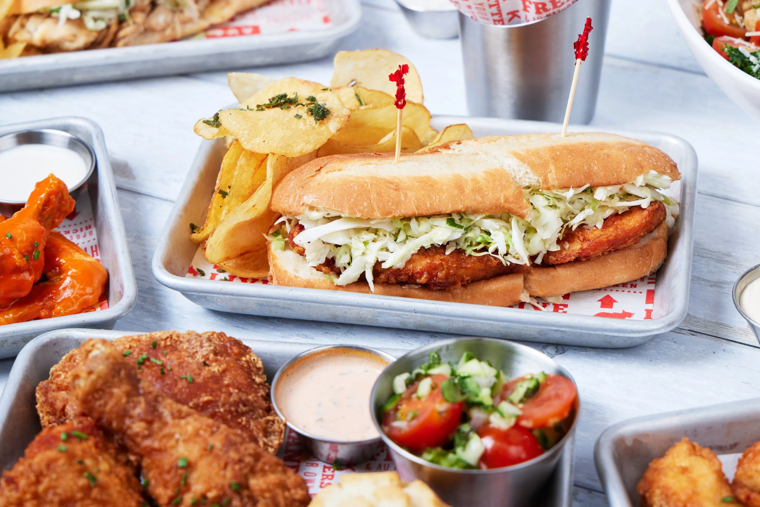 A close-up of a chicken sandwich with shredded lettuce in a bun, served with potato chips, surrounded by small bowls of tomato salad, fried chicken, and dipping sauces.