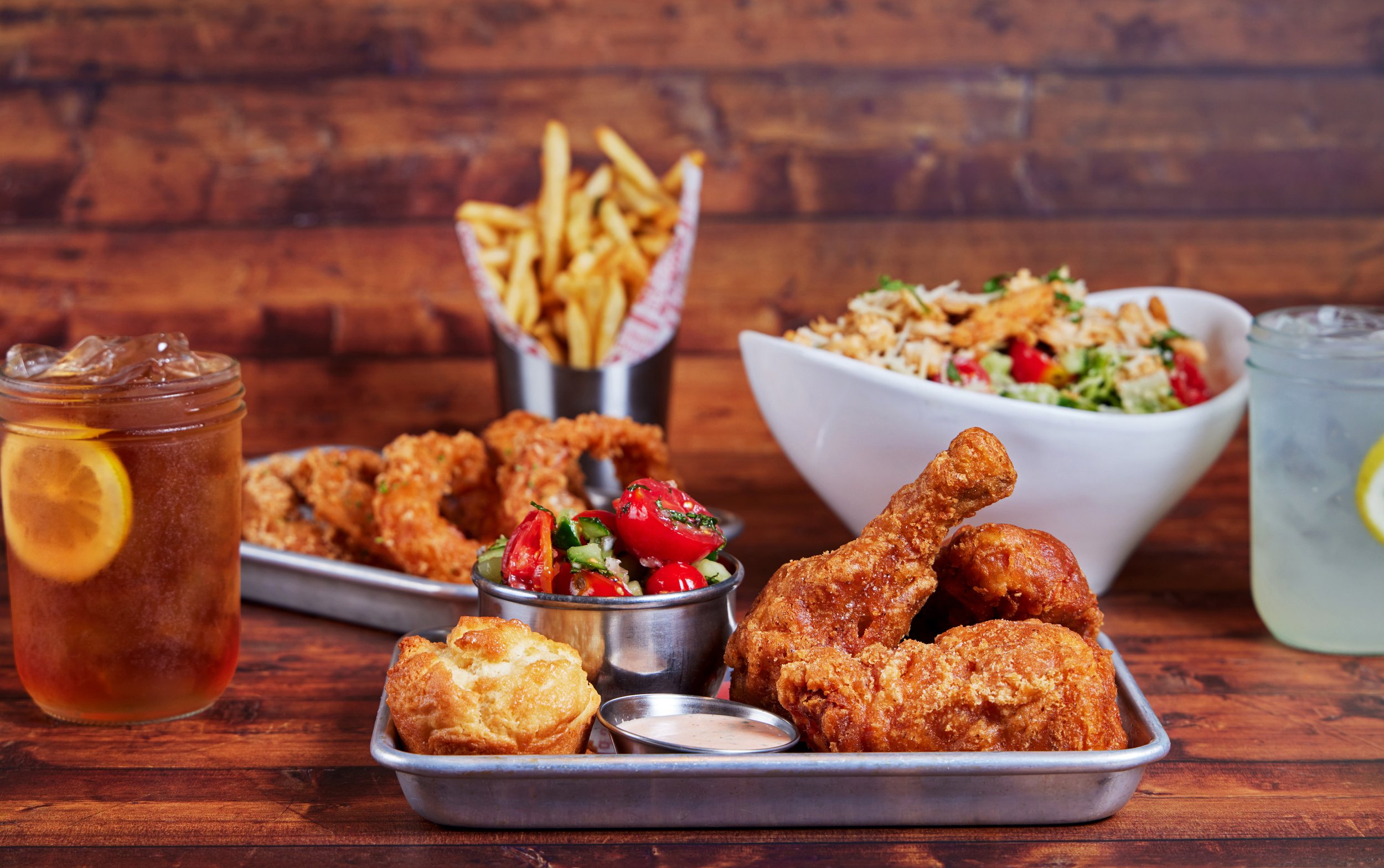 A tray of fried chicken pieces with honey mustard sauce, a cherry tomato salad, and onion rings, with iced tea and water on a wooden table.