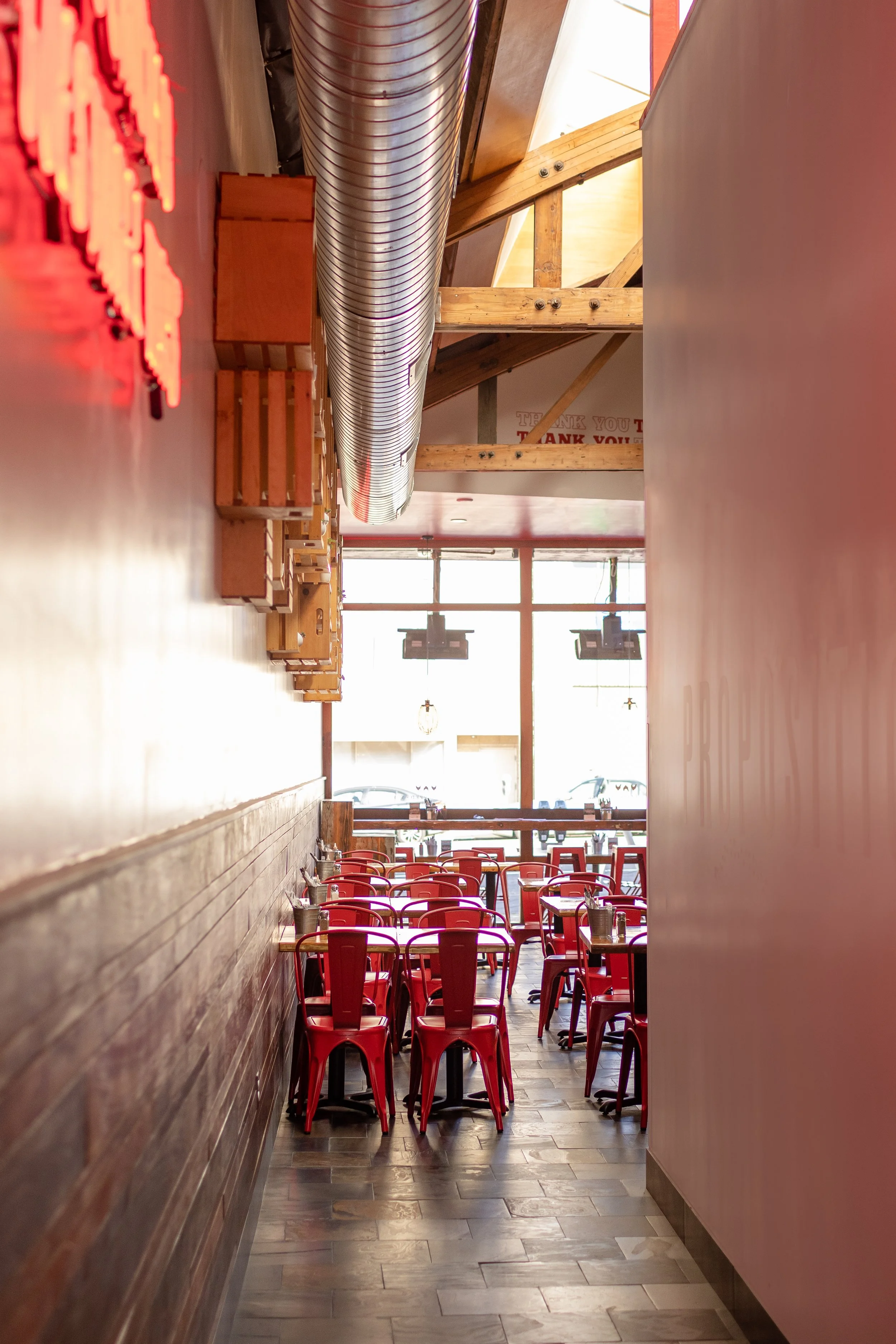 View down a hallway at Proposition Chicken in San Francisco, with wooden walls and red chairs, tables set with utensils, leading to a lit area with large windows.