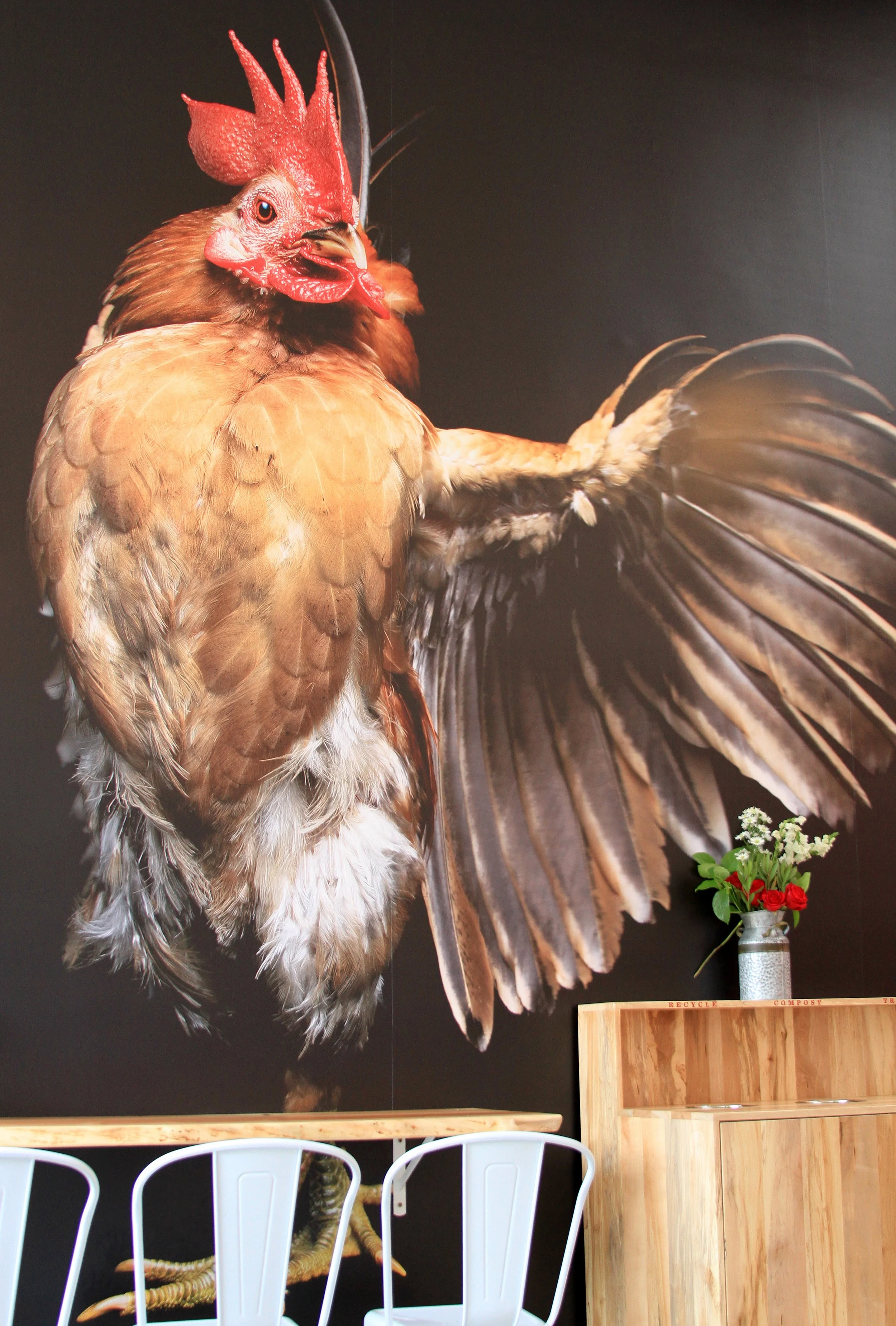 Interior of Proposition Chicken Oakland, featuring a large, detailed mural of a rooster with a dark background, a small table with four white chairs, and a wooden cabinet with a floral arrangement and greenery in a silver vase.