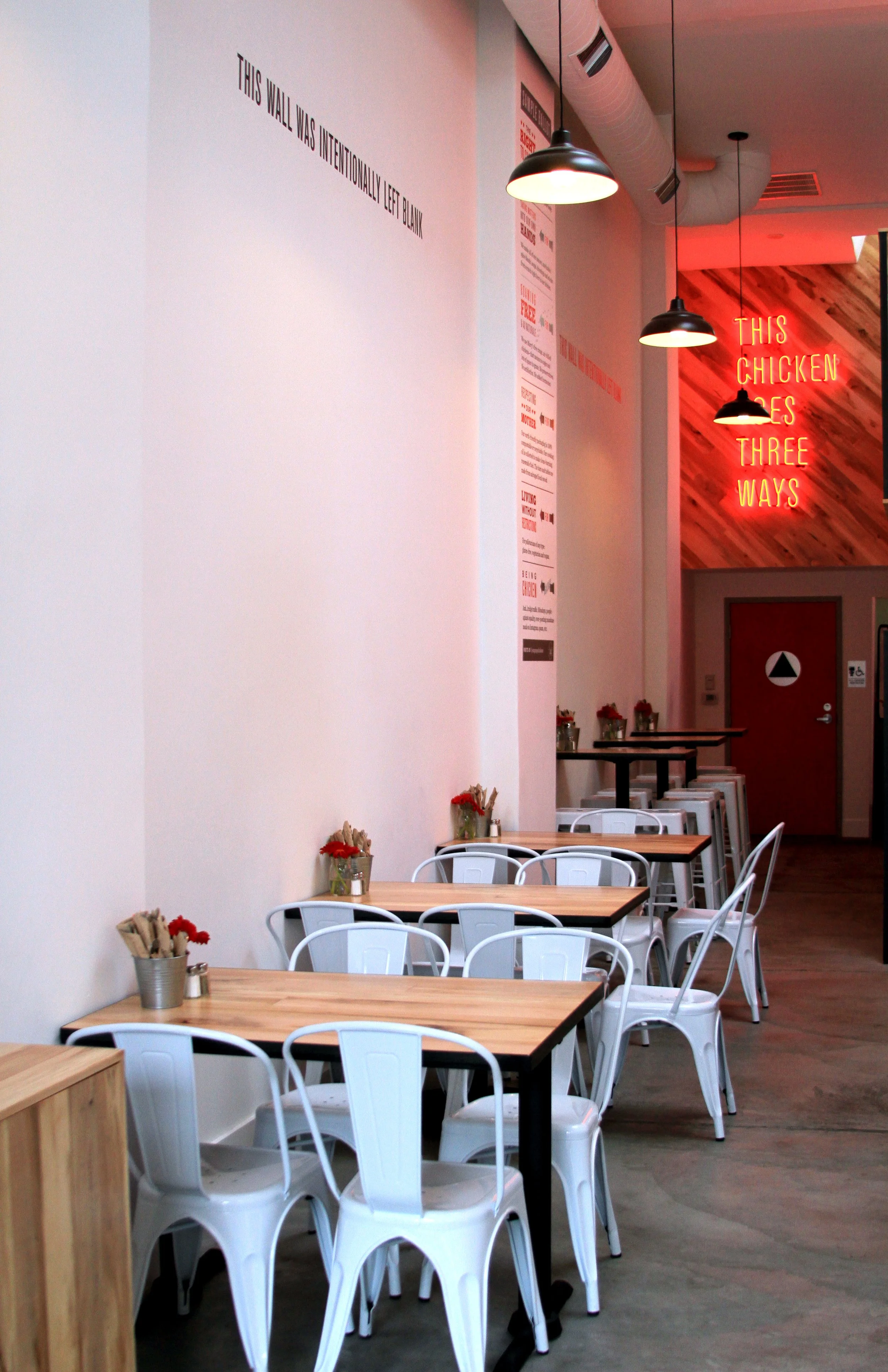 Inside Proposition Chicken Oakland seating area with tables and chairs. There are bright lighting fixtures hanging from the ceiling. A neon sign on a wooden accent wall displays the words 'This chicken goes three ways.'