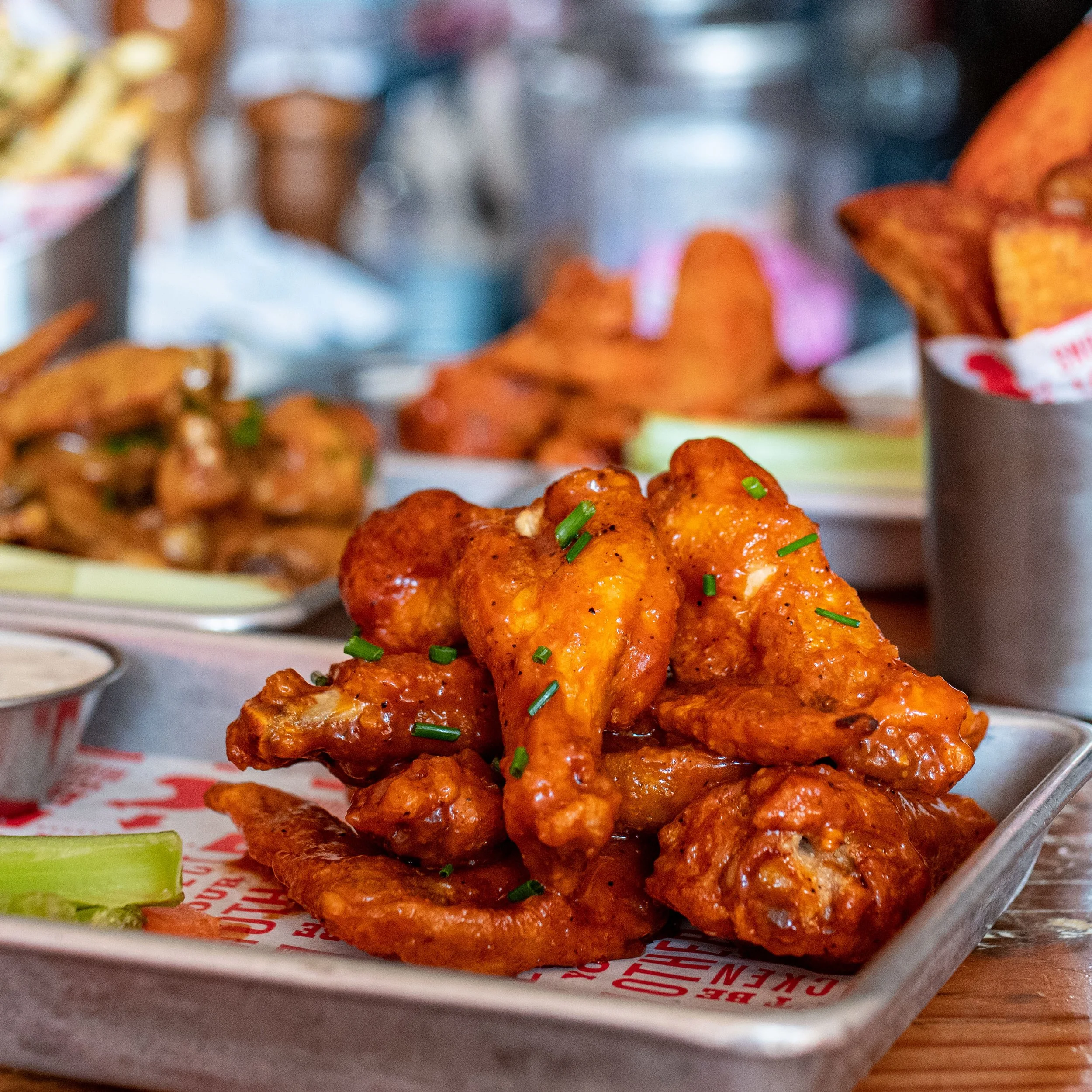 Close-up of chicken wings garnished with chopped green onions, on a tray lined with red and white paper, with another tray of chicken wings and a container of celery sticks partially visible in the background.