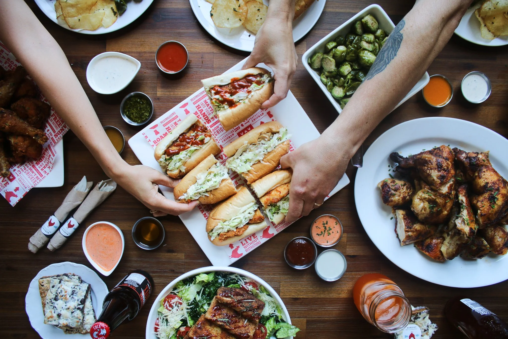 A variety of dishes including sandwiches, chicken wings, salads, fries, and condiments on a wooden table at a casual meal setting.