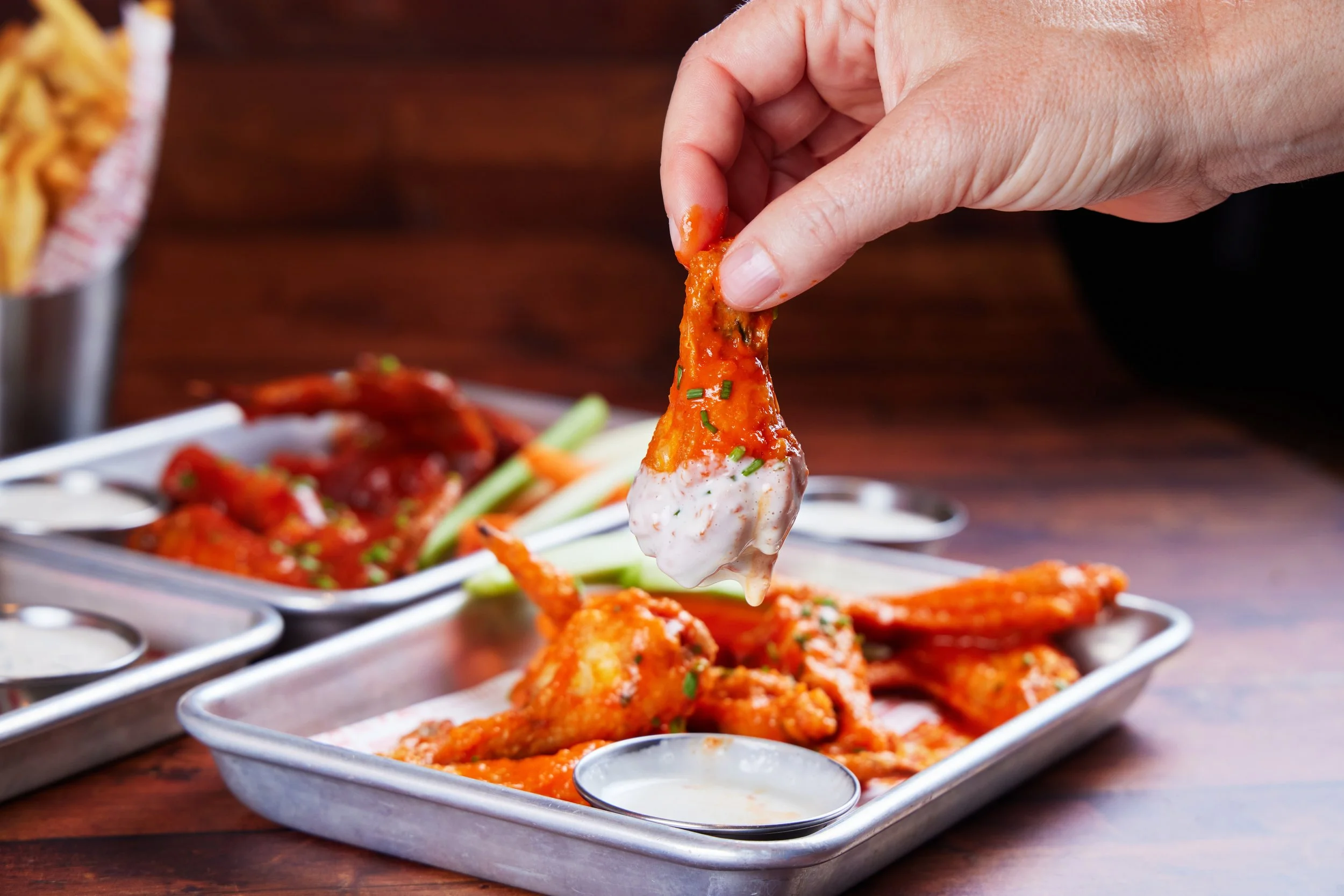 Close-up of a hand picking up a piece of buffalo chicken wing coated in sauce with a small bowl of ranch or blue cheese dip in the foreground, behind a tray of buffalo chicken wings garnished with chopped herbs.