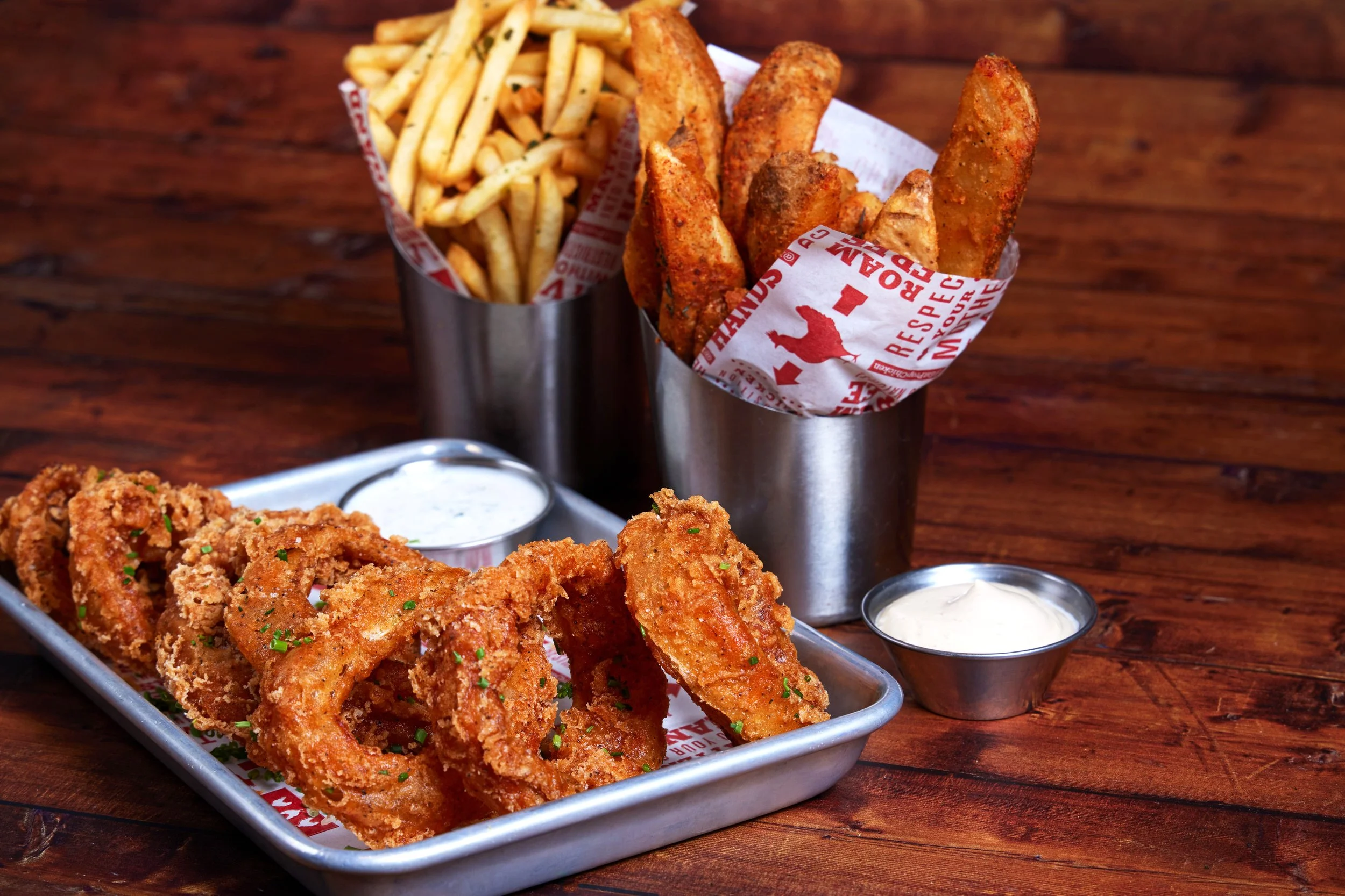 Fried onion rings on a metal tray with chopped parsley, served with small cups of ranch dipping sauce, alongside a metal cup of French fries, on a wooden table.