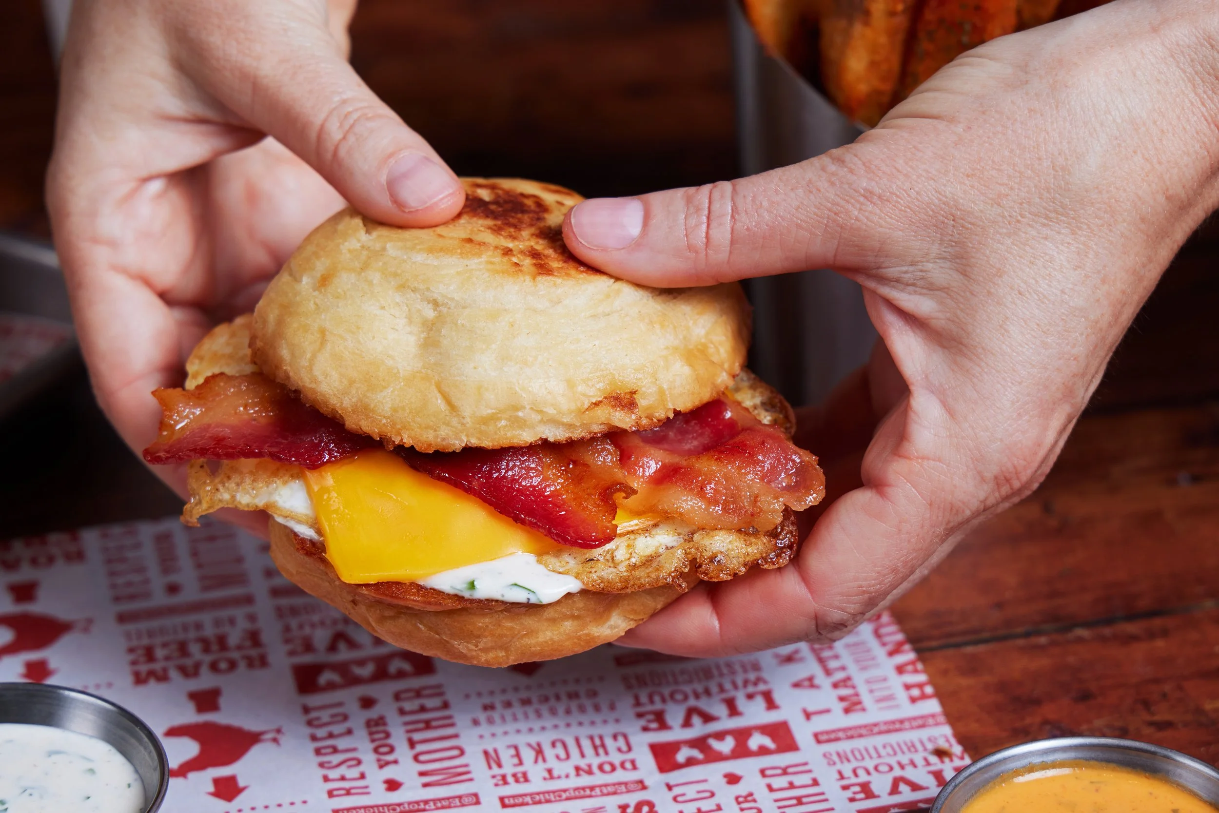 Close-up of a person using their fingers to hold and assemble a breakfast sandwich with fried egg, chicken, cheddar cheese, bacon, and bun. There are condiments in metal cups on a red and white paper-lined surface.