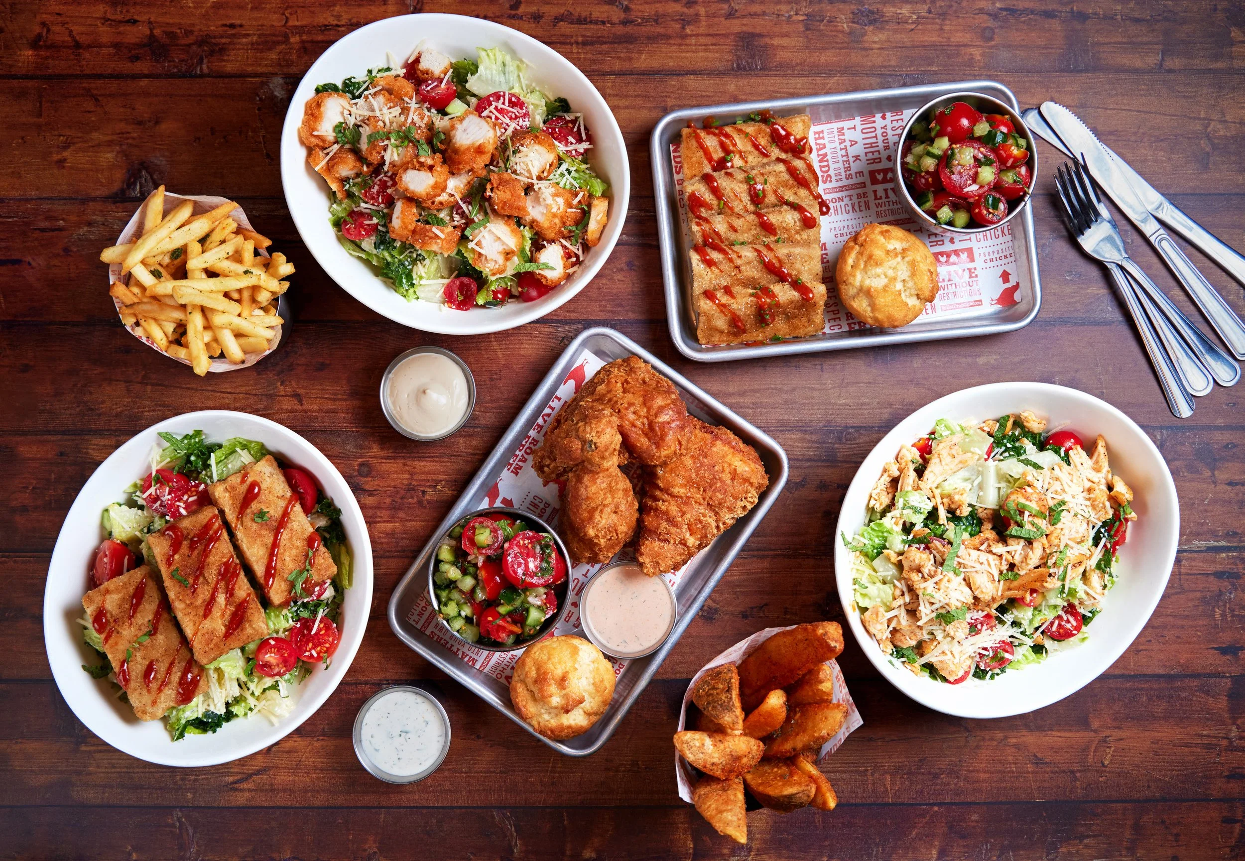 Assorted dishes of fried chicken, salads, French fries, potato wedges, and dipping sauces on a wooden table.