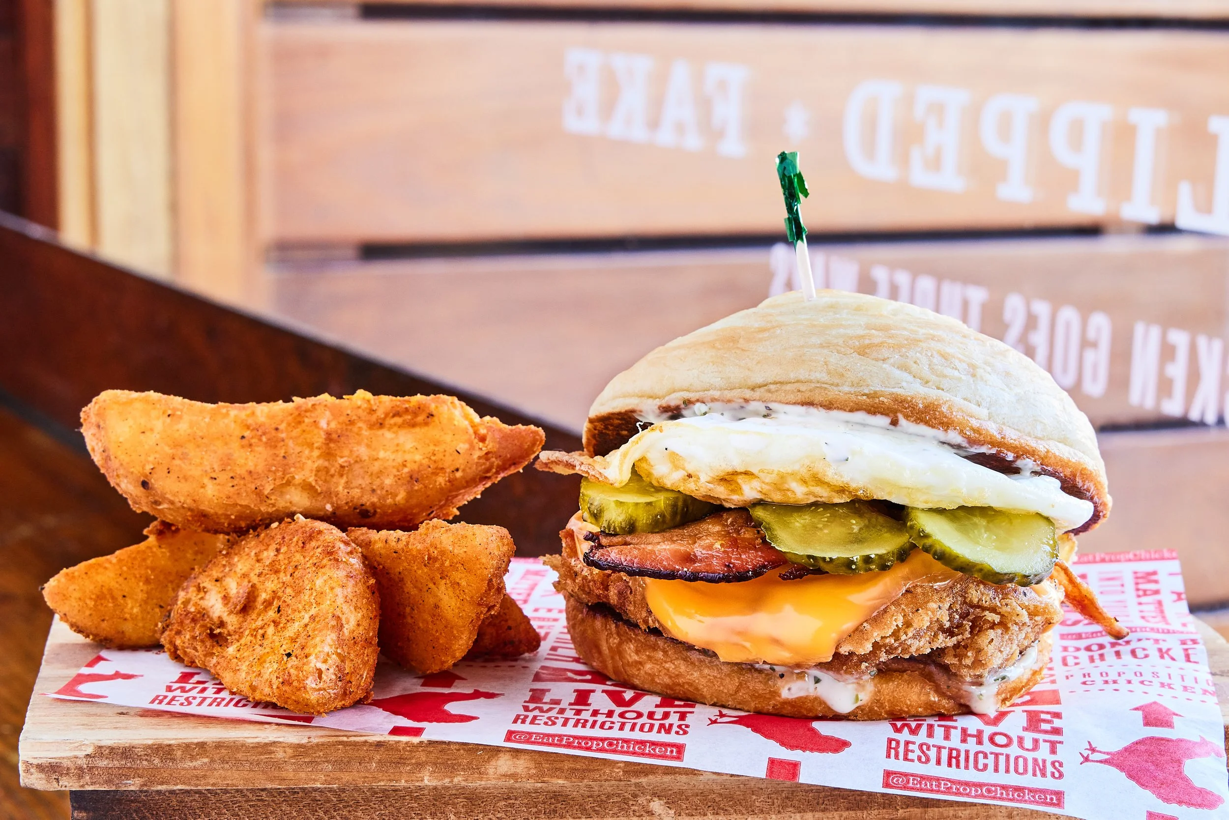 Close-up of a fried chicken and egg breakfast sandwich on a toasted bun, served with seasoned potato wedges on red and white checkered paper.