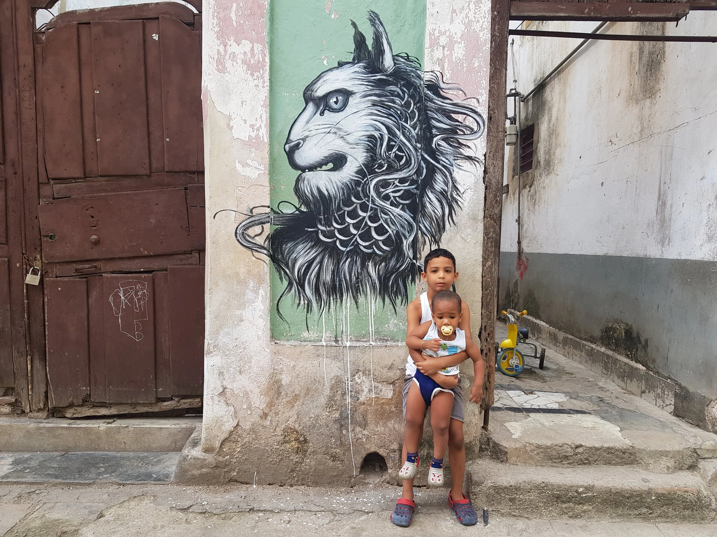Two young children, a boy holding a baby, standing in front of a street wall with graffiti art of a lion's head. The lion is depicted in black and white with detailed mane and piercing eyes. The boy holds a pacifier and is looking directly at the cam