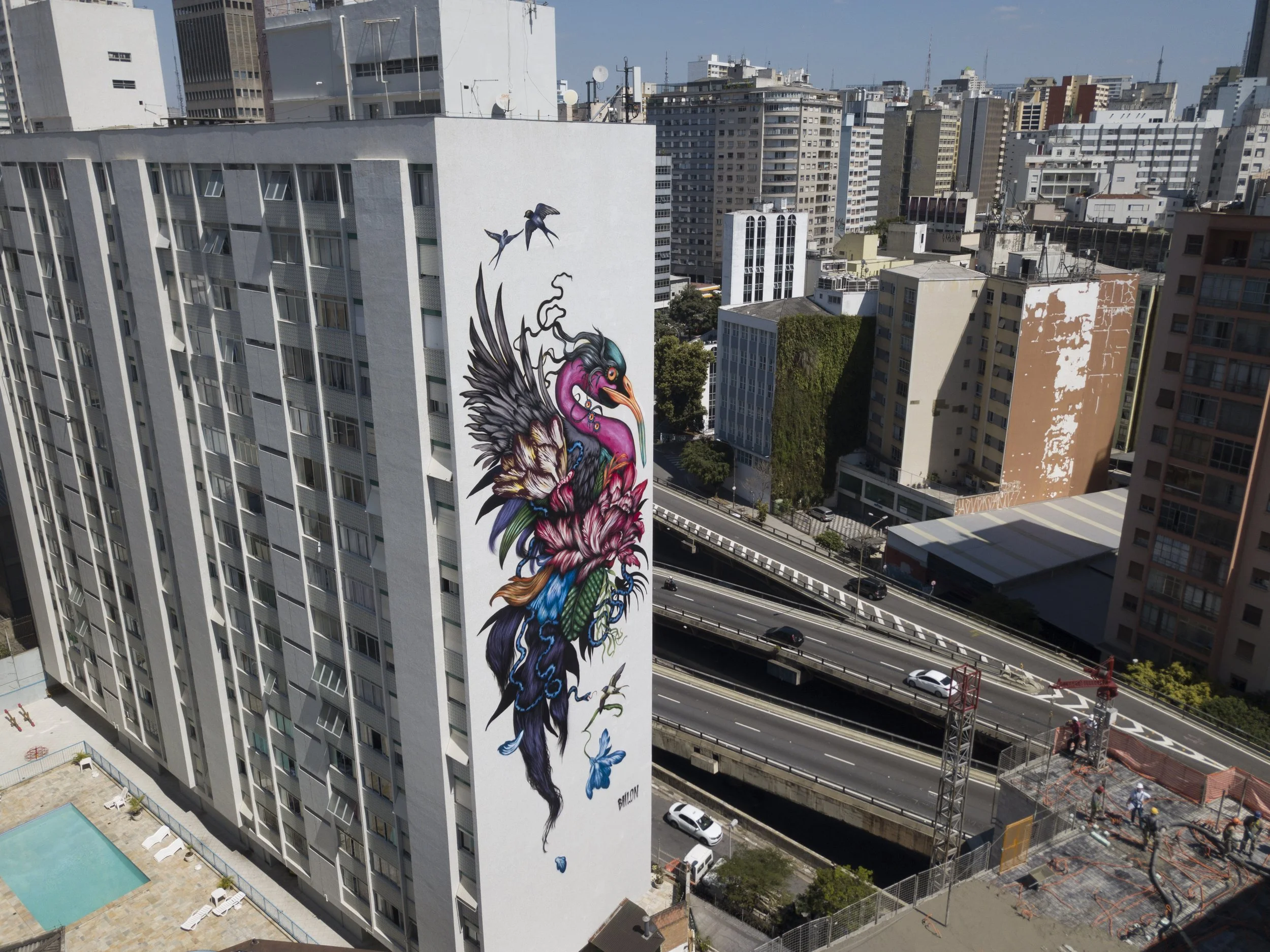A tall building in an urban area with a large, colorful mural of a bird with vibrant feathers and flowers painted on its side. The cityscape includes several other high-rise buildings, a street with moving cars, and a construction site nearby.