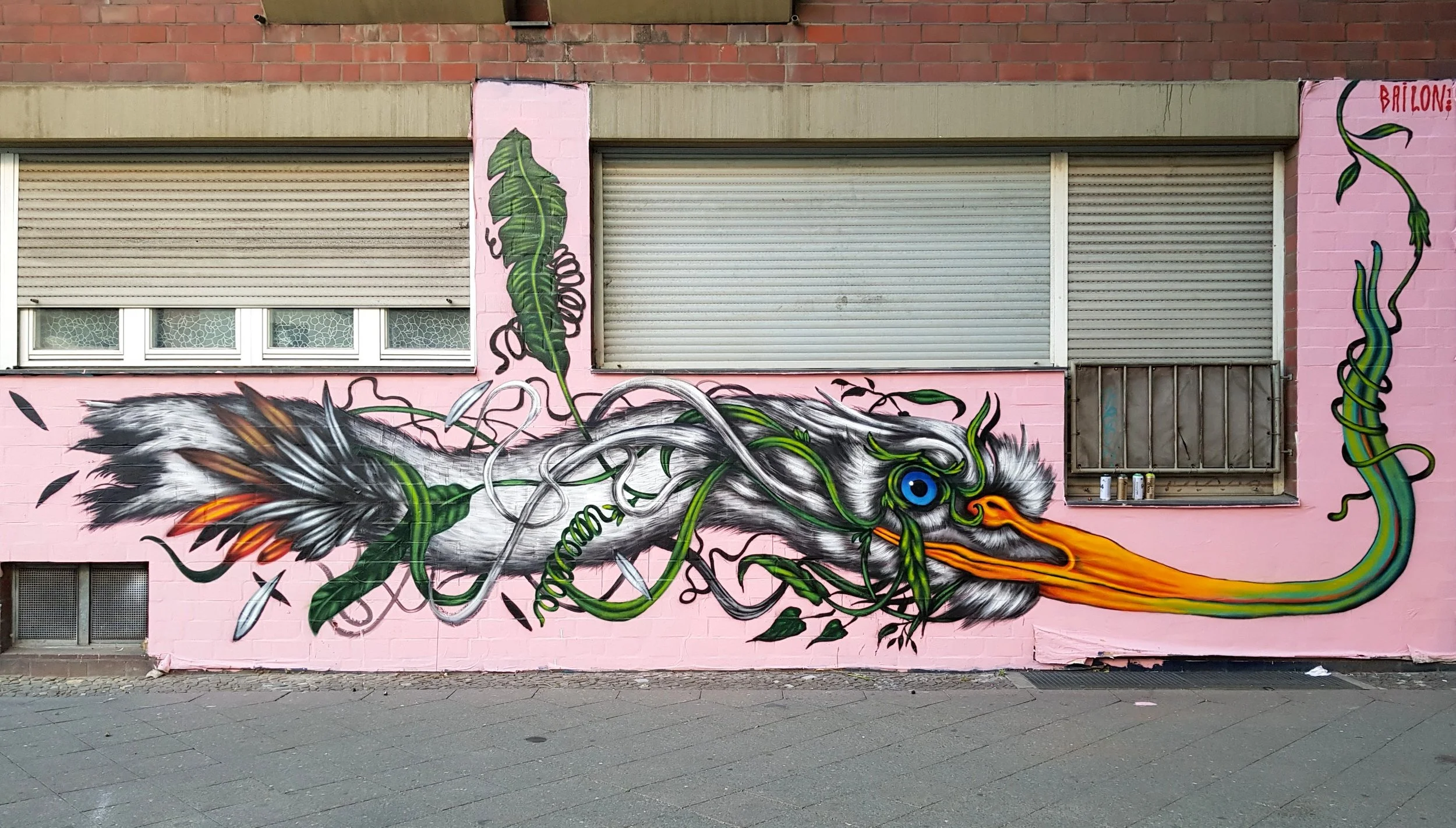 Colorful graffiti mural of a bird with an exaggerated long beak and large blue eye, surrounded by green and black vines, painted on a pink wall with graffiti-style details.