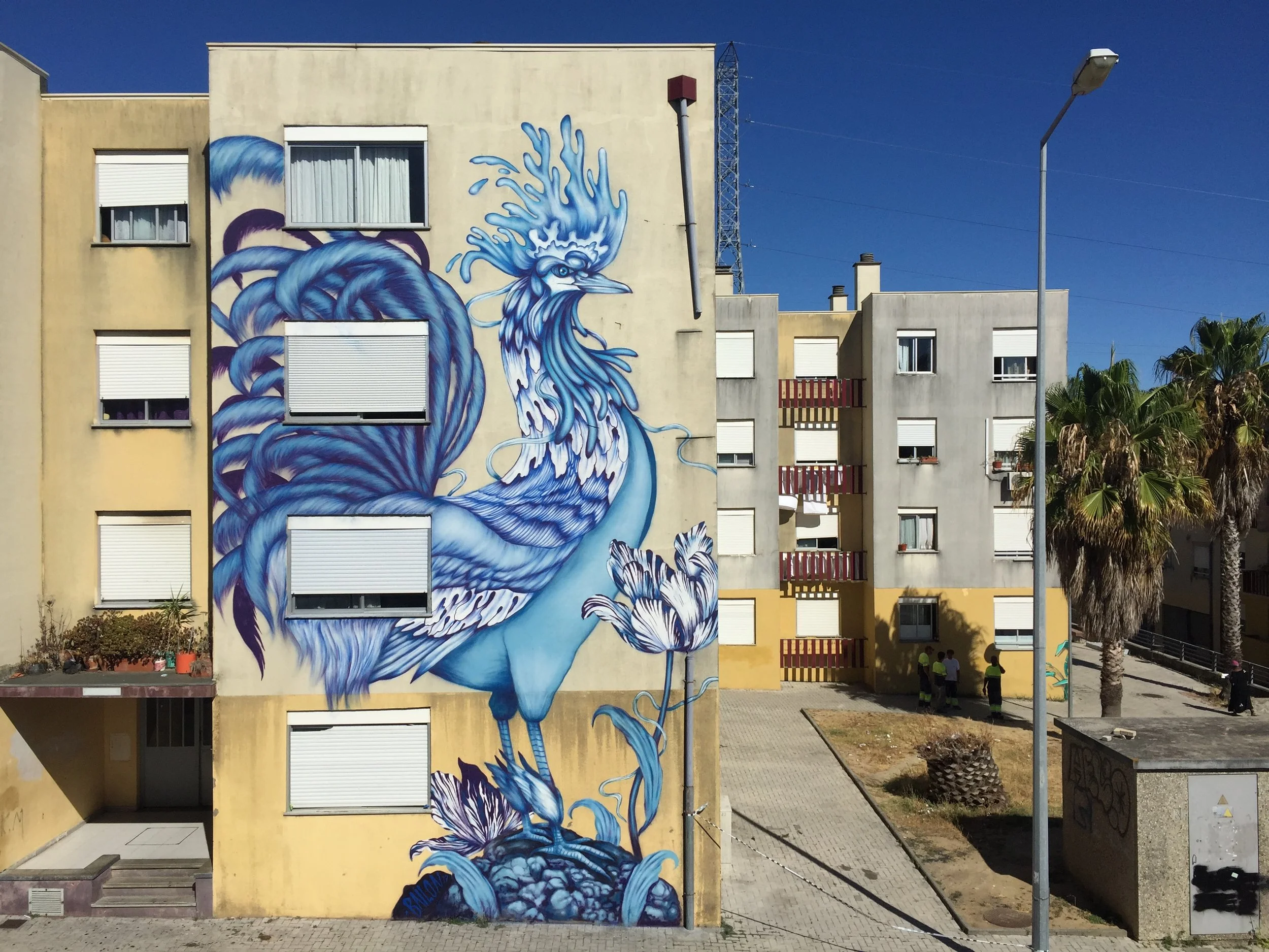 A large mural of a blue and white stylized phoenix on the side of a beige apartment building with multiple windows and closed white blinds. Streetlamps, palm trees, and some people are visible nearby under a clear blue sky.