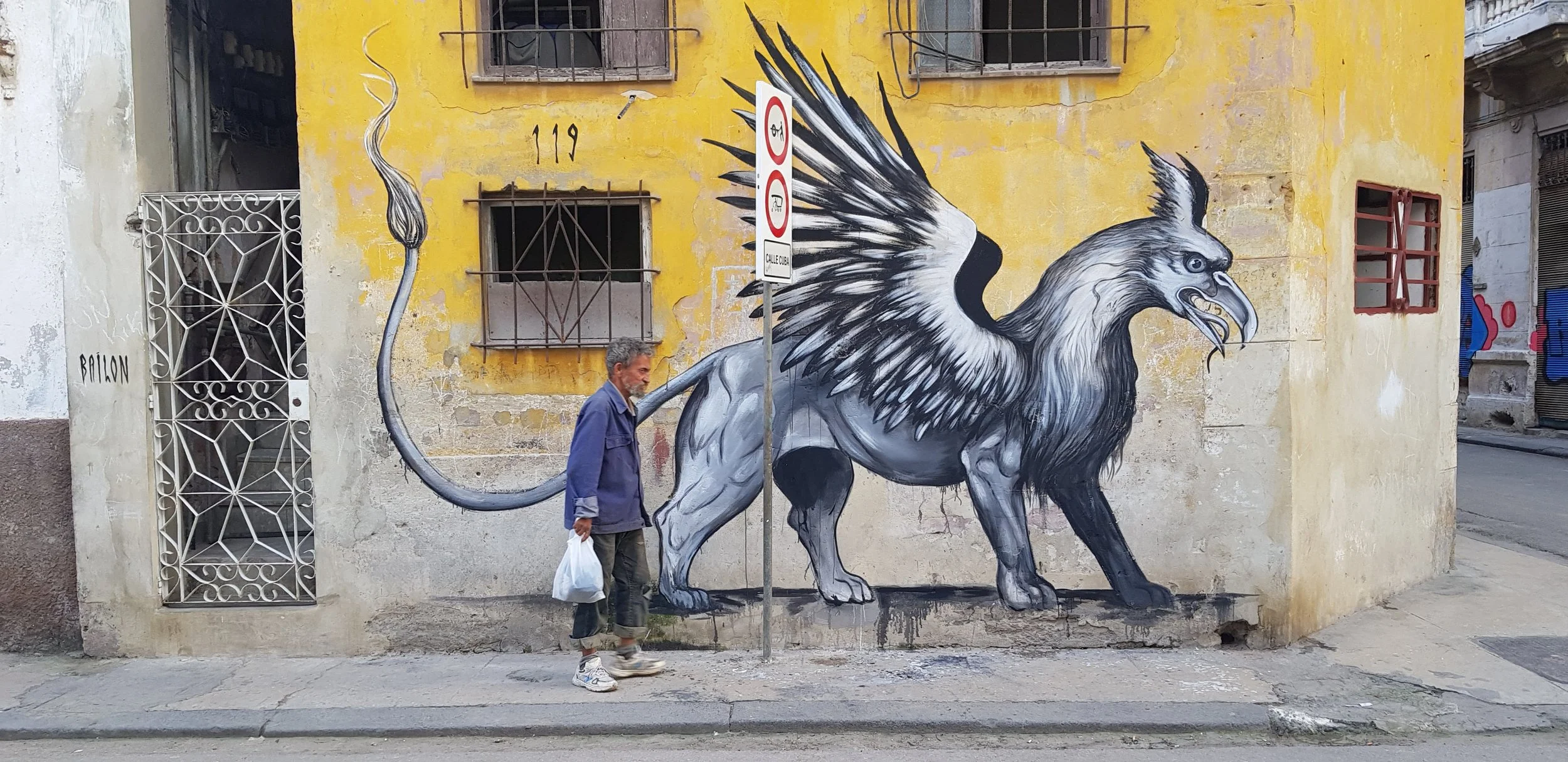 Street art mural of a mythical creature with the body of a lion and the head of an eagle on a yellow building wall, with a man walking past and a street sign in front.