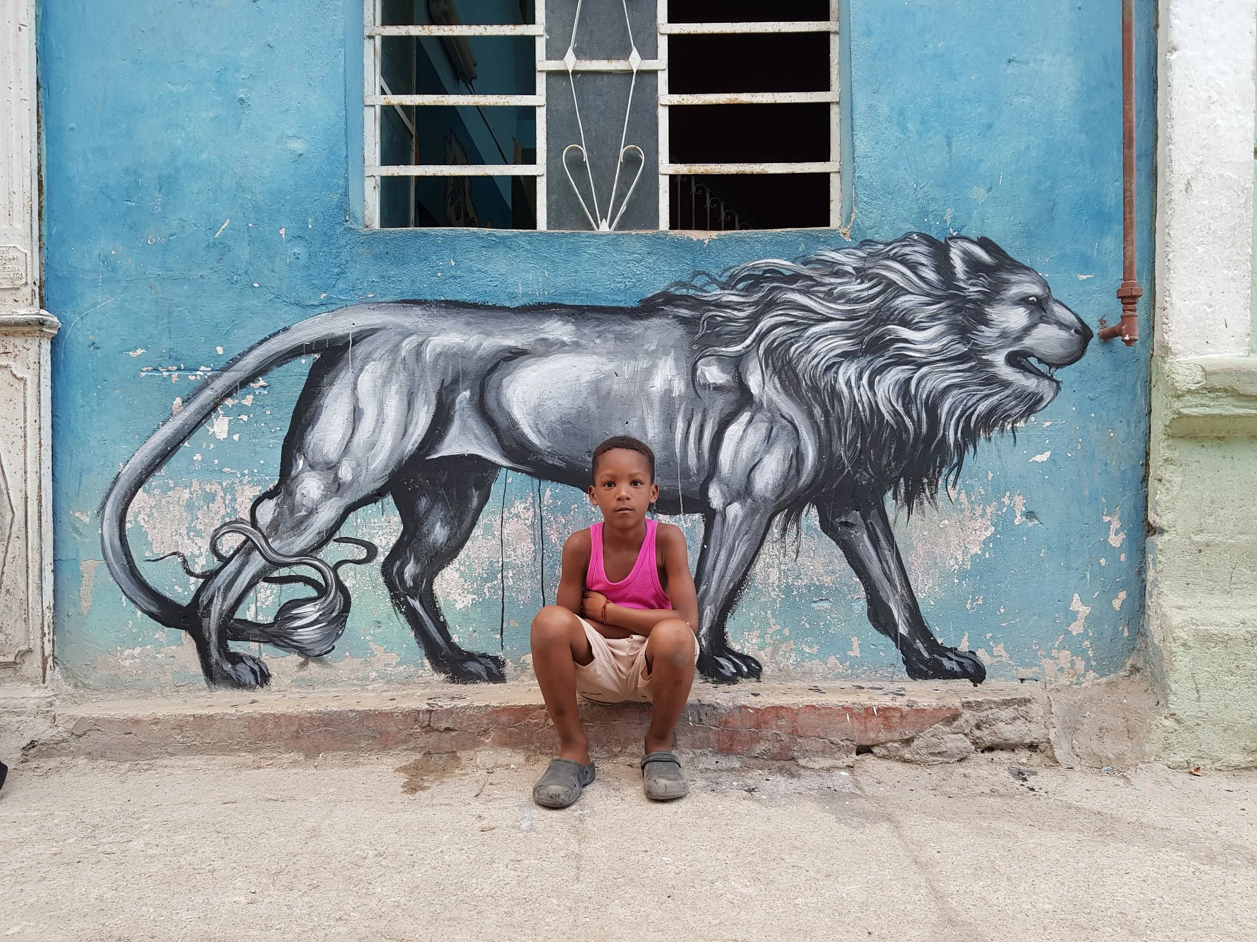 A young boy wearing a pink tank top, shorts, and sandals sits on a step in front of a mural of a black and gray lion painted on a blue wall. The boy is looking directly at the camera with a neutral expression.