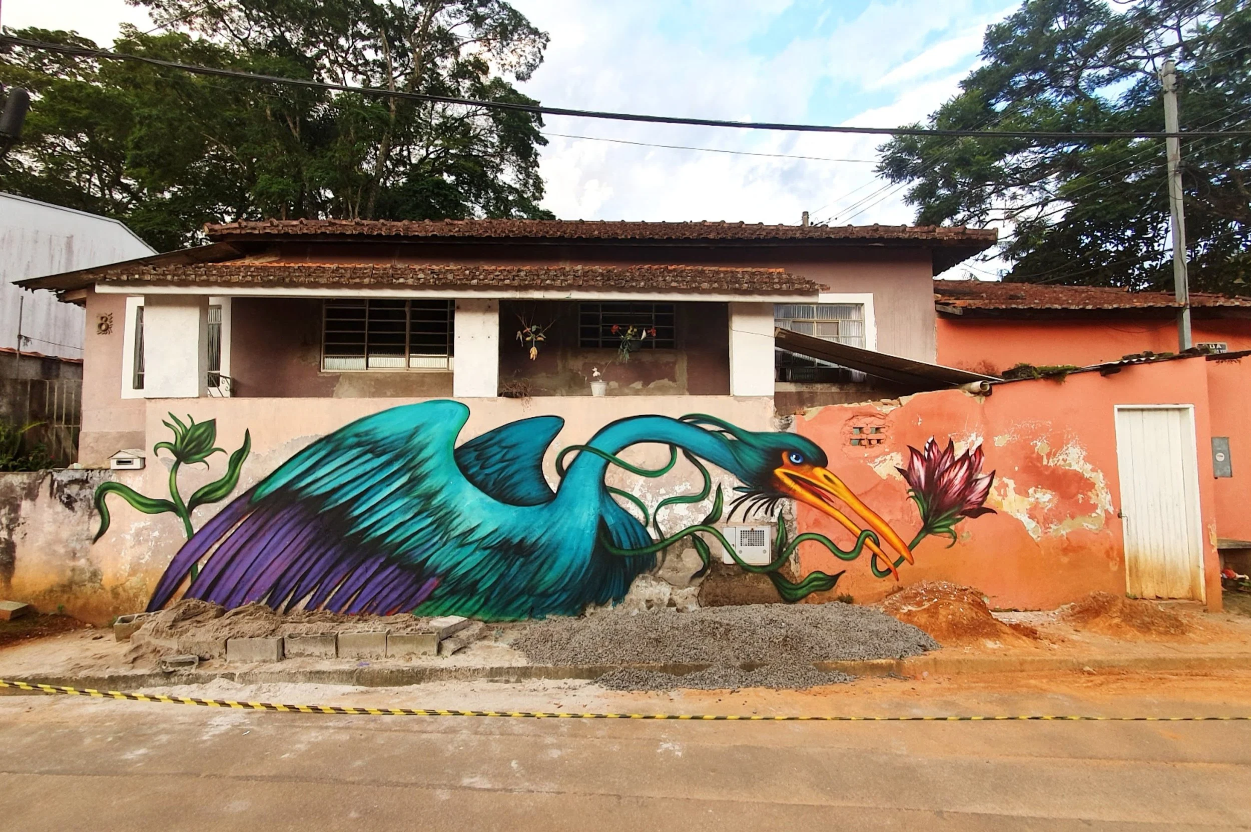 Colorful mural of a blue heron on a pink building wall, with flowers and greenery in the background.