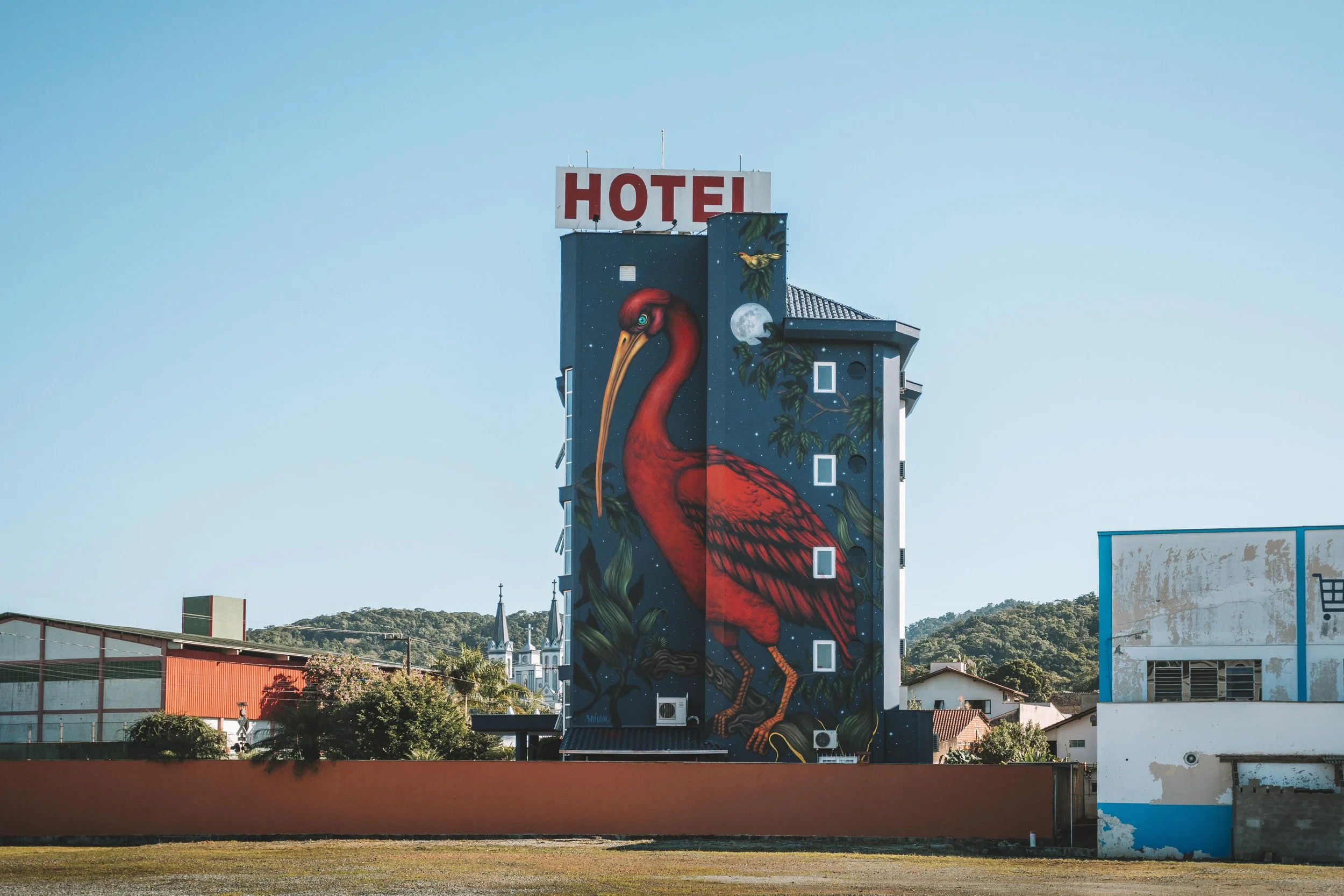 A tall building features a large mural of a red flamingo with a detailed background, including trees, leaves, a full moon, and small birds. The building has a red and white hotel sign on top and small windows along the mural. The surrounding area inc