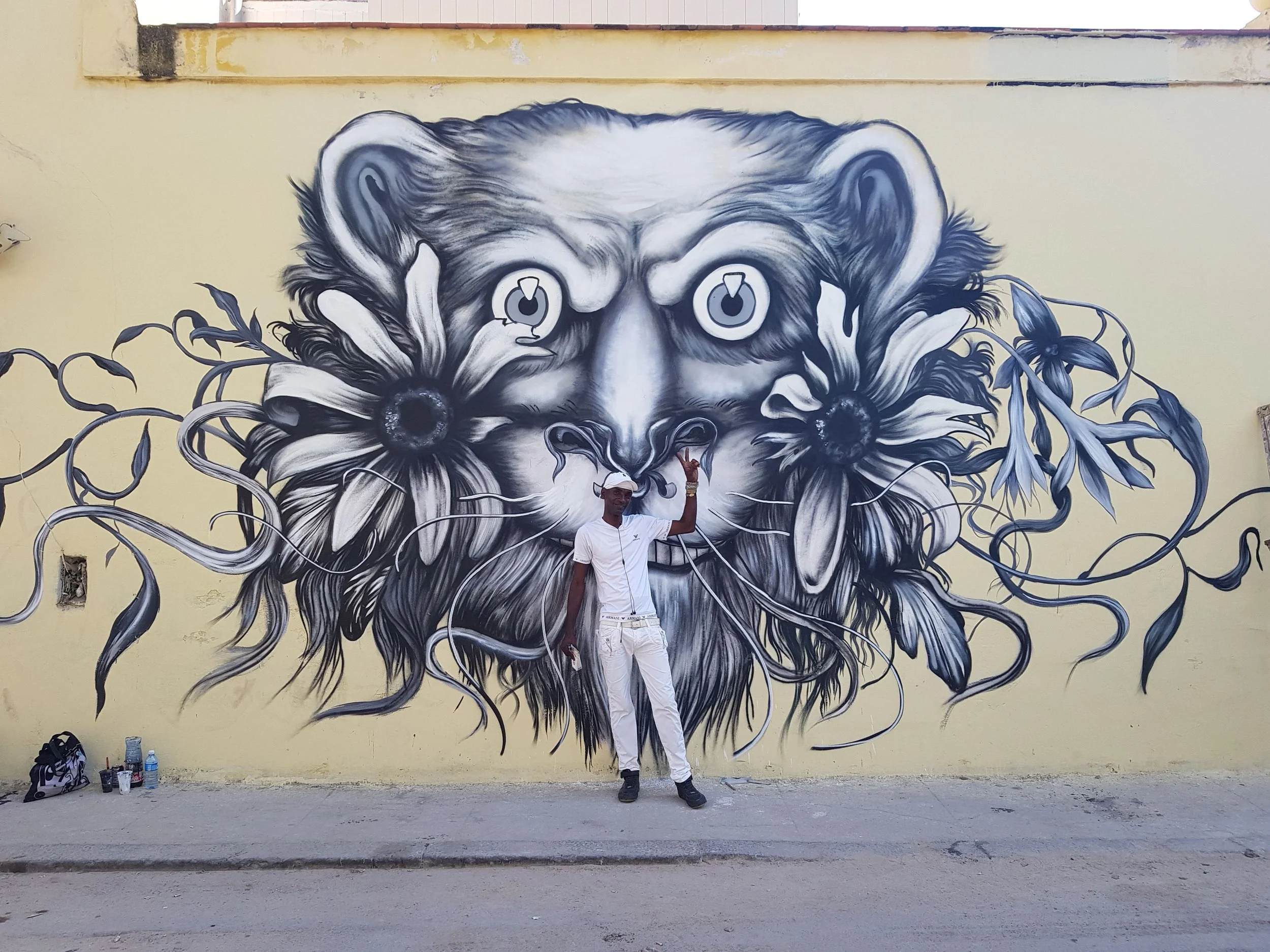 A man in white clothing and a white cap poses in front of a large, monochromatic mural of a lion's face painted on a yellow wall. The man is standing near the center with one hand raised pointing to the sky. Art supplies and water bottles are on the 