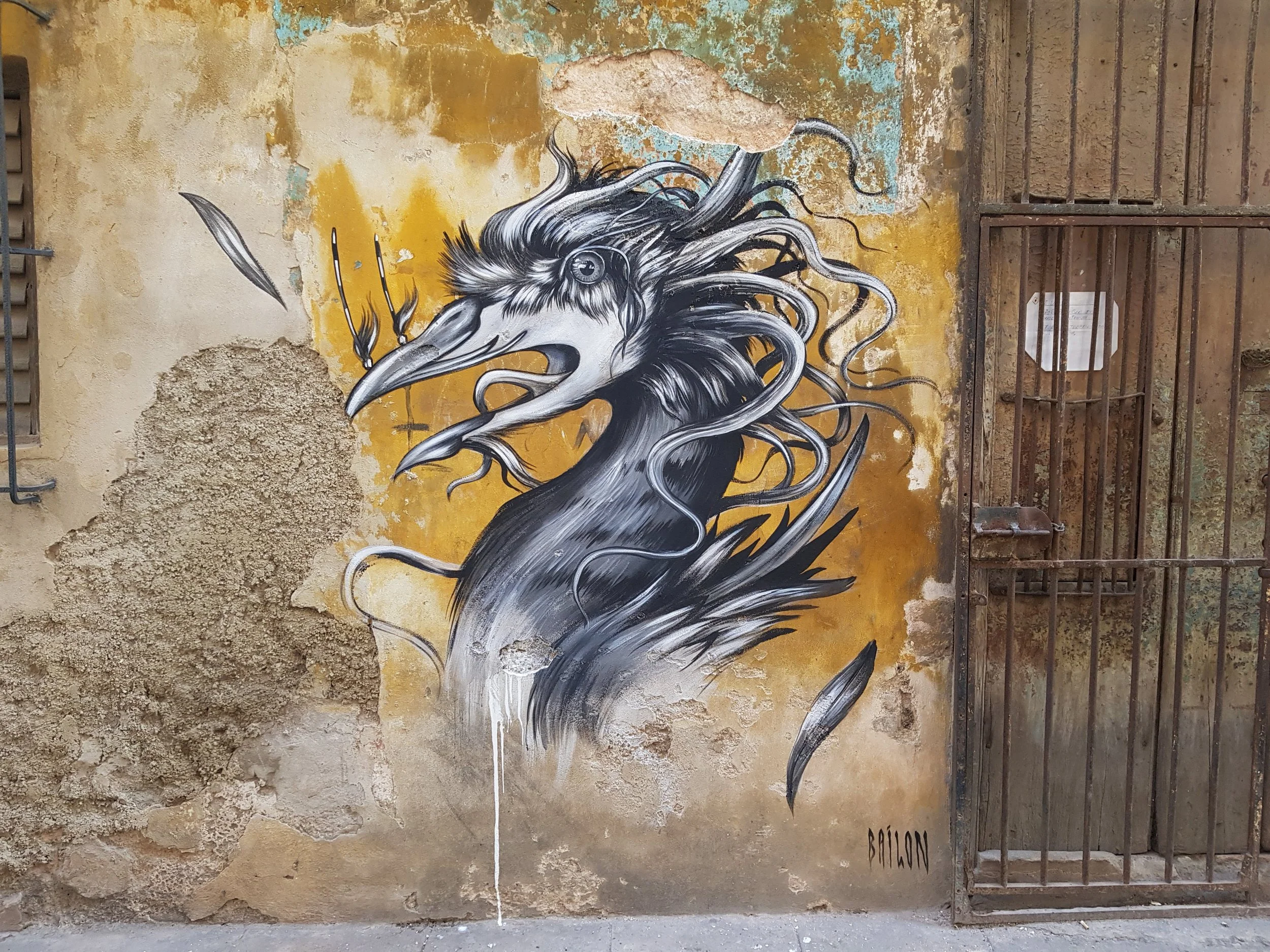 Street art mural of a black and white horse with flowing mane painted on an old, distressed wall with peeling paint and a rusted gate.