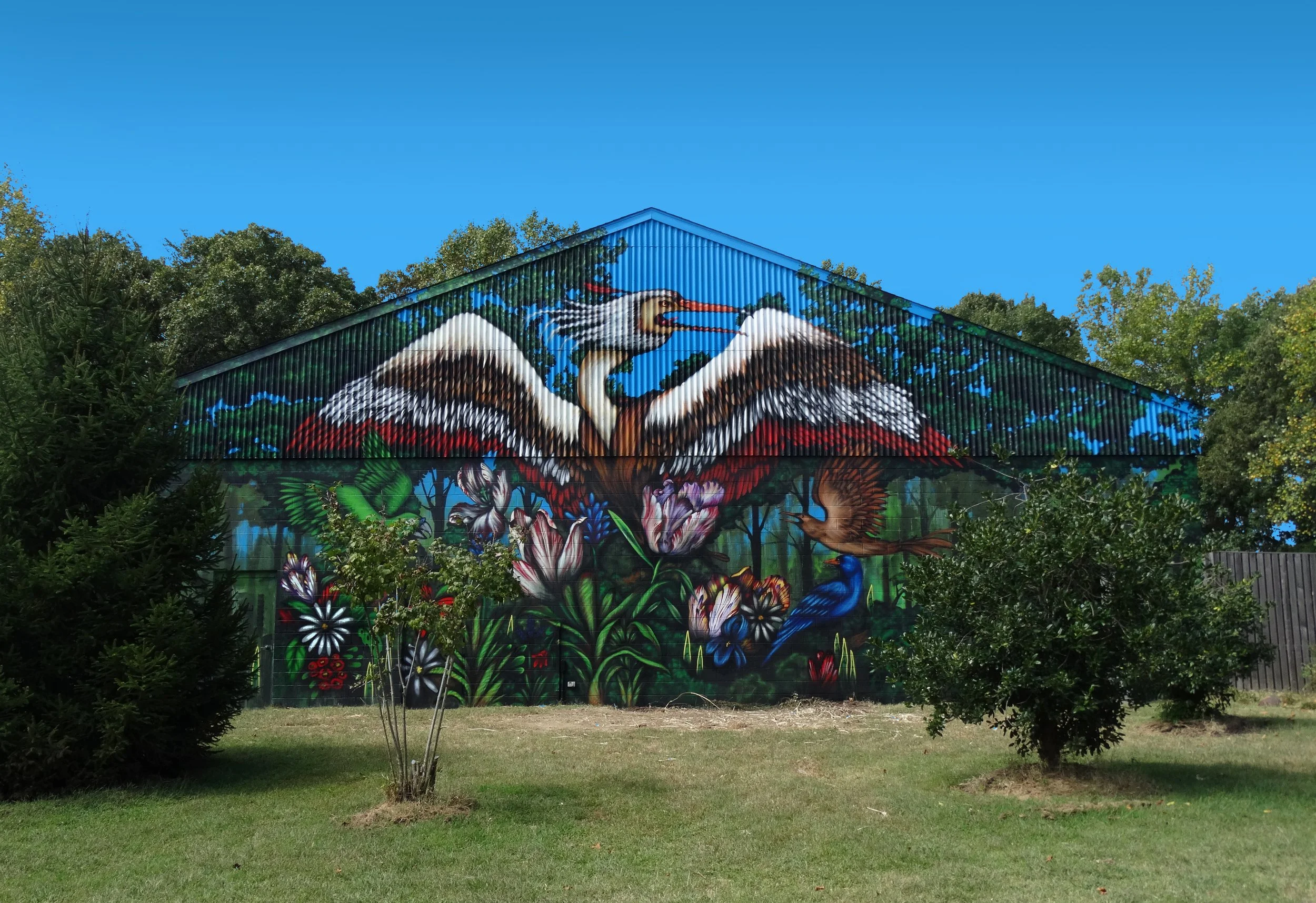 Colorful mural of two storks surrounded by lush greenery and bright flowers on a building wall, with a clear blue sky above.