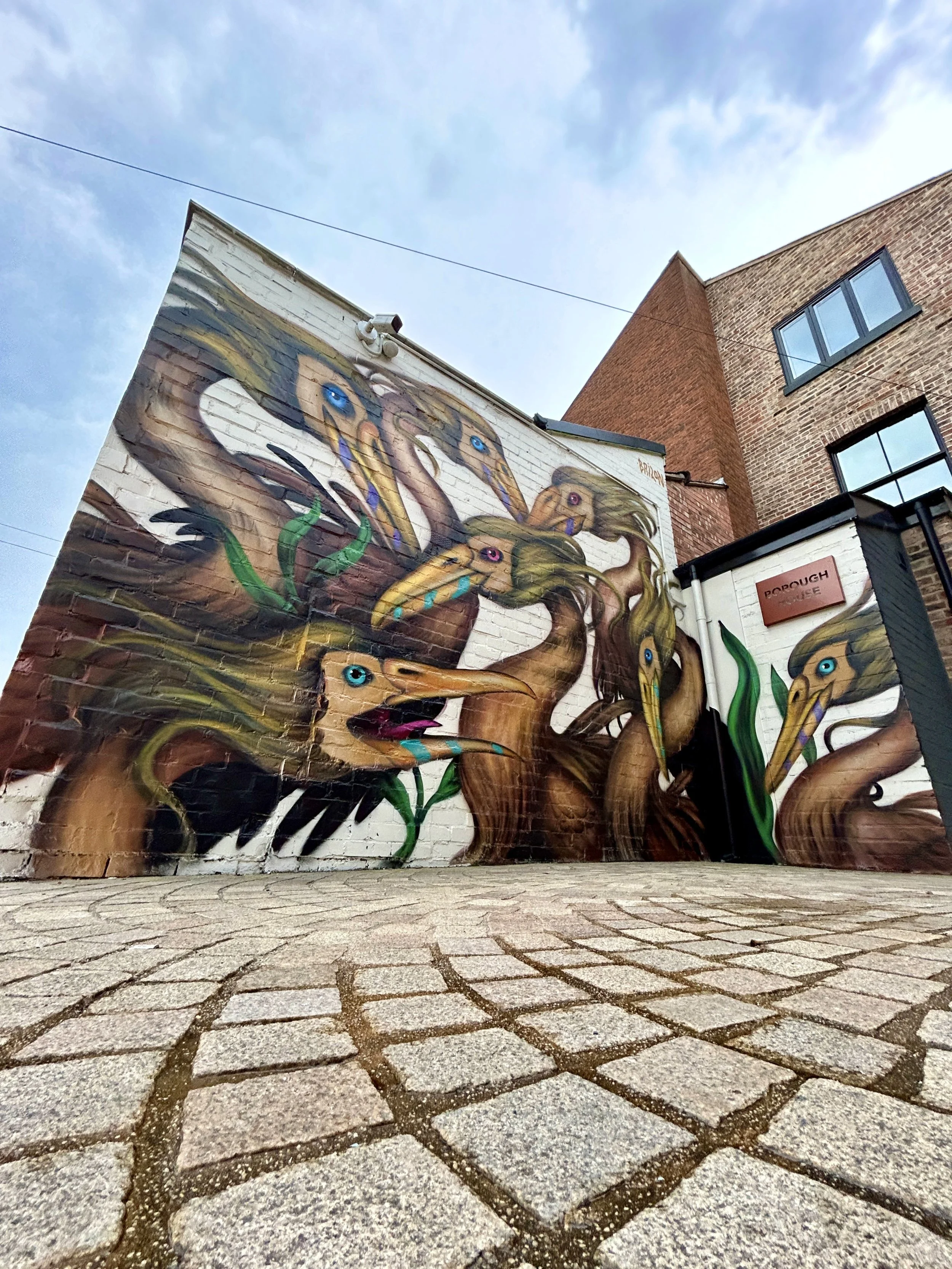 Colorful mural painting of mermaids with blue eyes and flowing hair on brick wall, under a cloudy sky, located in an urban setting.