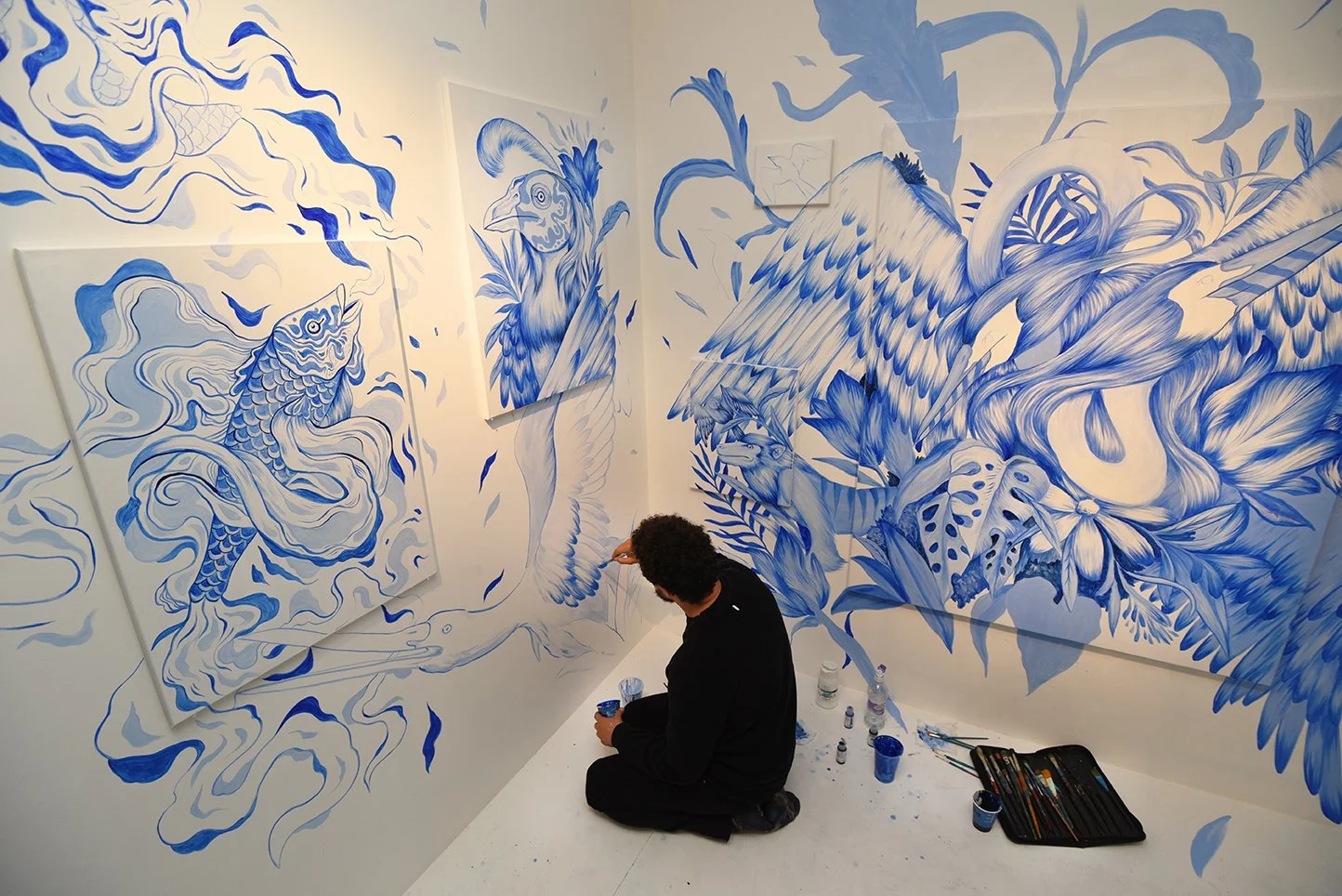 An artist painting detailed blue and white murals on the walls and canvases, featuring aquatic and nature-inspired imagery.