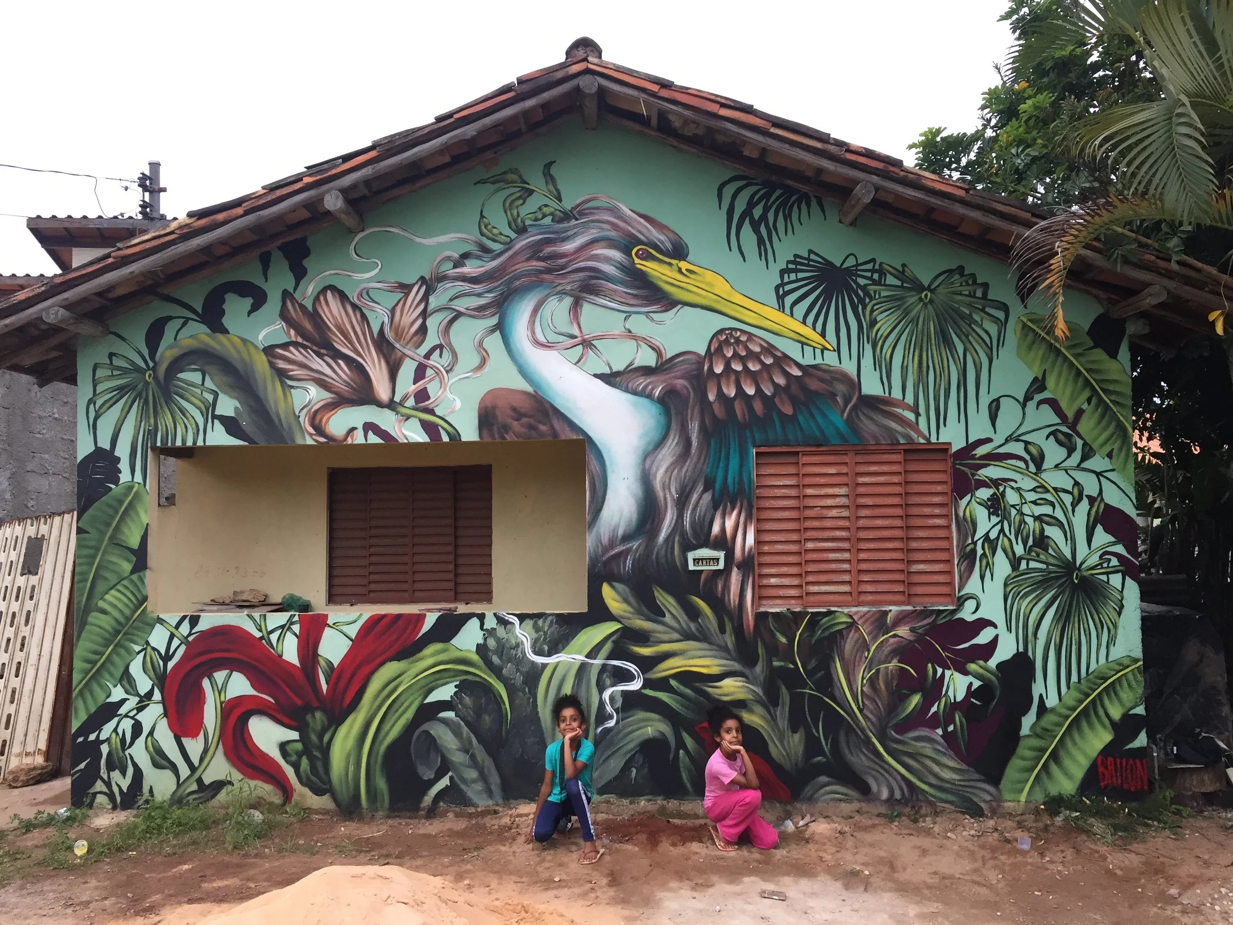 A mural of a heron with a long beak and flowing feathers is painted on the side of a building, surrounded by lush green plants and leaves. Two children are crouching in front of the mural.
