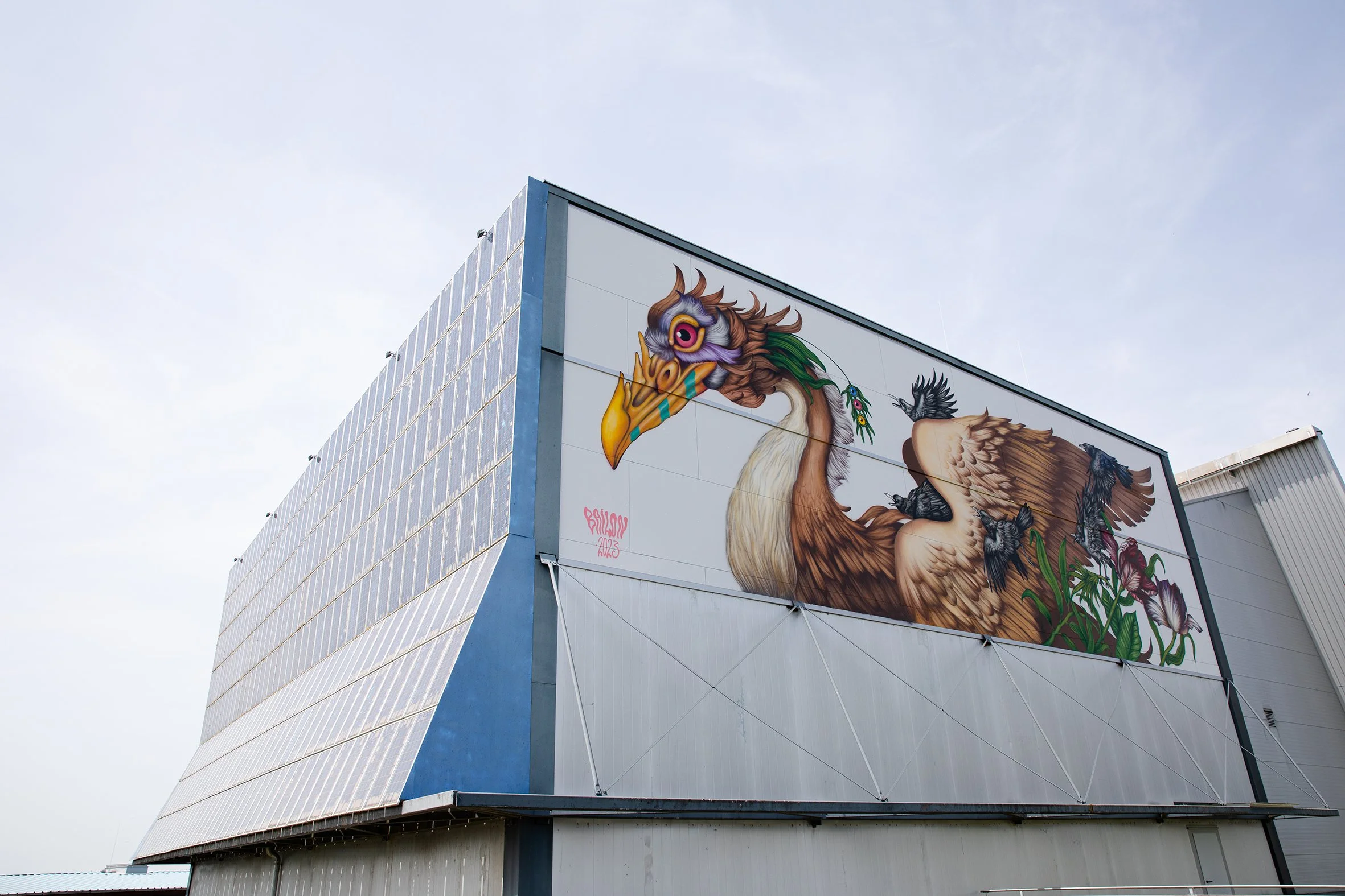 A large mural of a mythological creature on the side of a modern building with solar panels. The creature is a colorful griffin with a bird's head and wings, and a lion's body, standing in a natural setting.