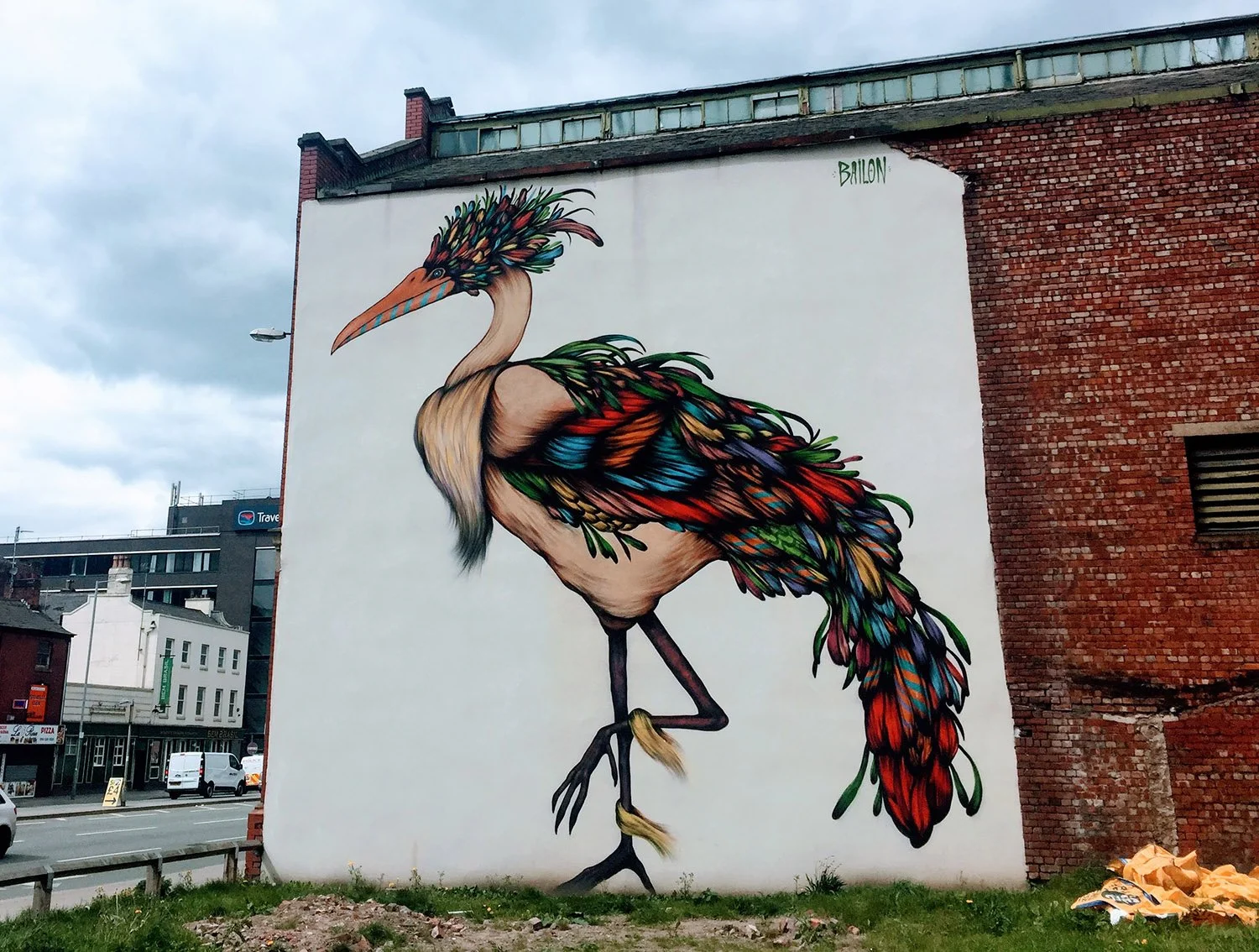 Large mural of a colorful, artistic bird on the side of a brick building, with a city street and cloudy sky in the background.