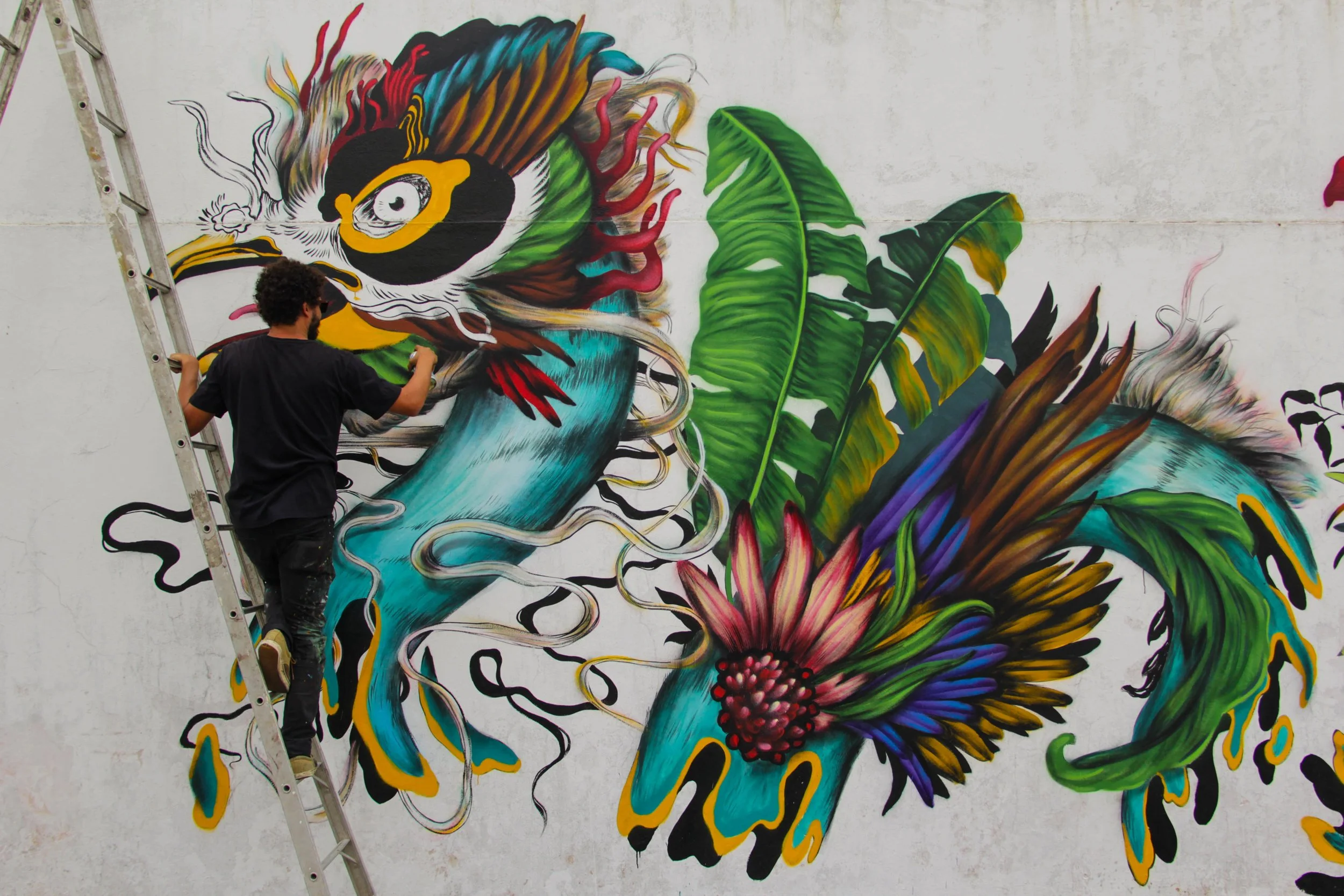 A man painting a colorful mural of a bird and vibrant flowers on a white wall.