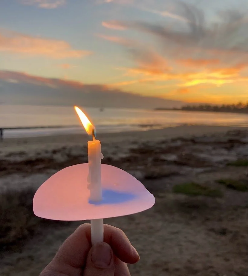 A hand holding a lit candle in front of a beach during sunset with colorful sky and calm ocean in the background.